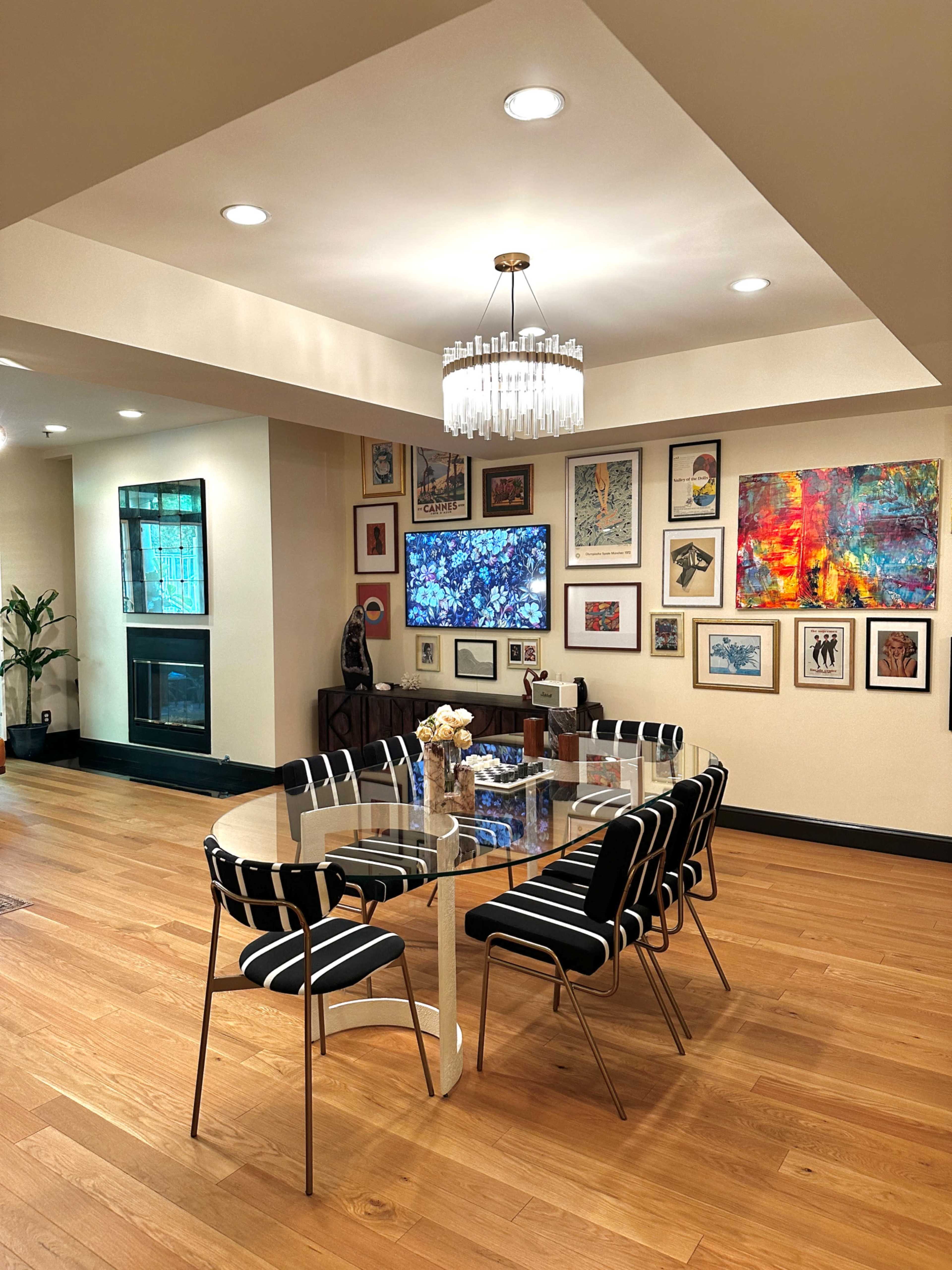 A dining area features a glass table surrounded by striped chairs, with a gallery wall of framed artwork and a fireplace.