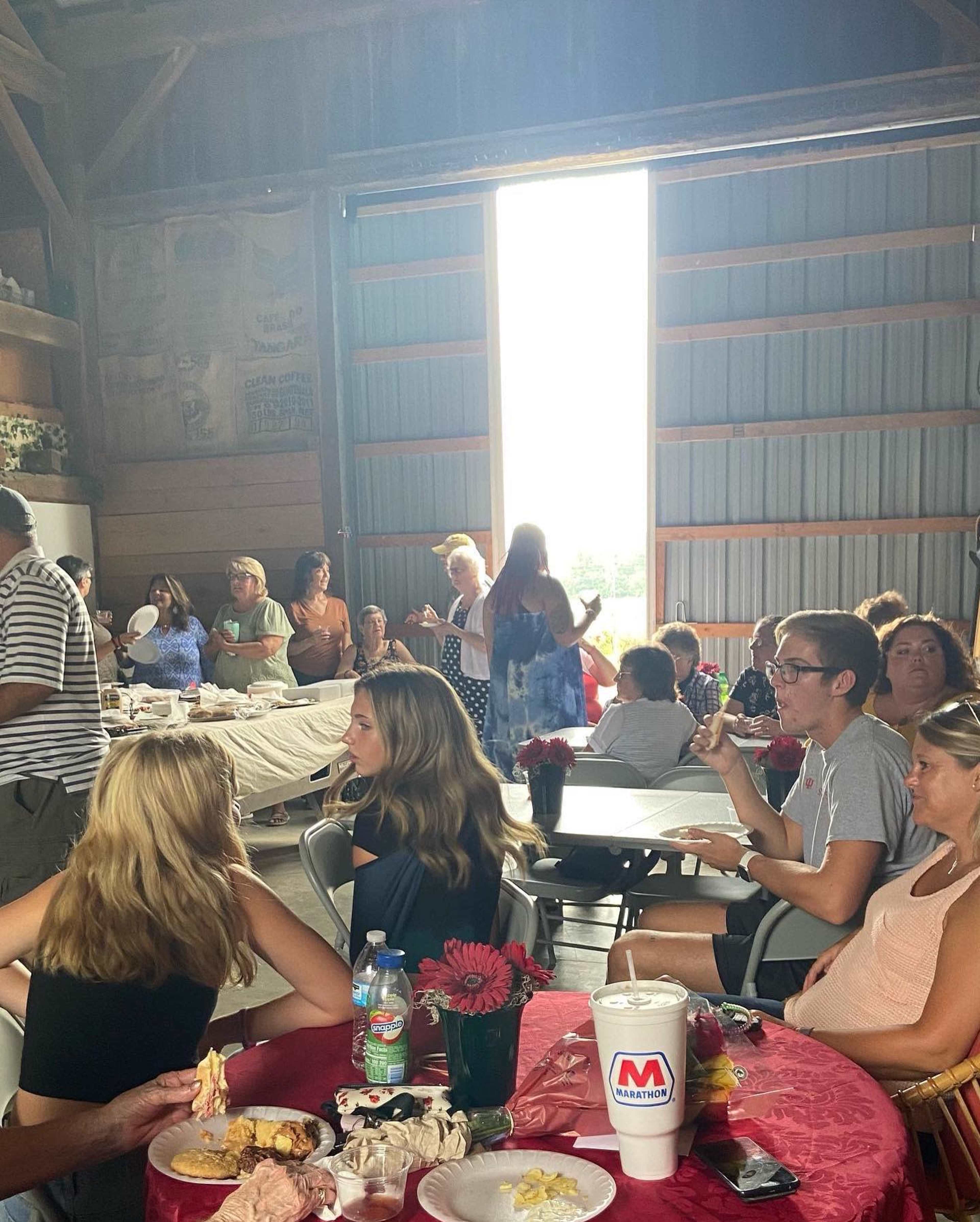 A group of people sit at tables inside a large barn, with sunlight streaming through an open door while others stand and socialize.