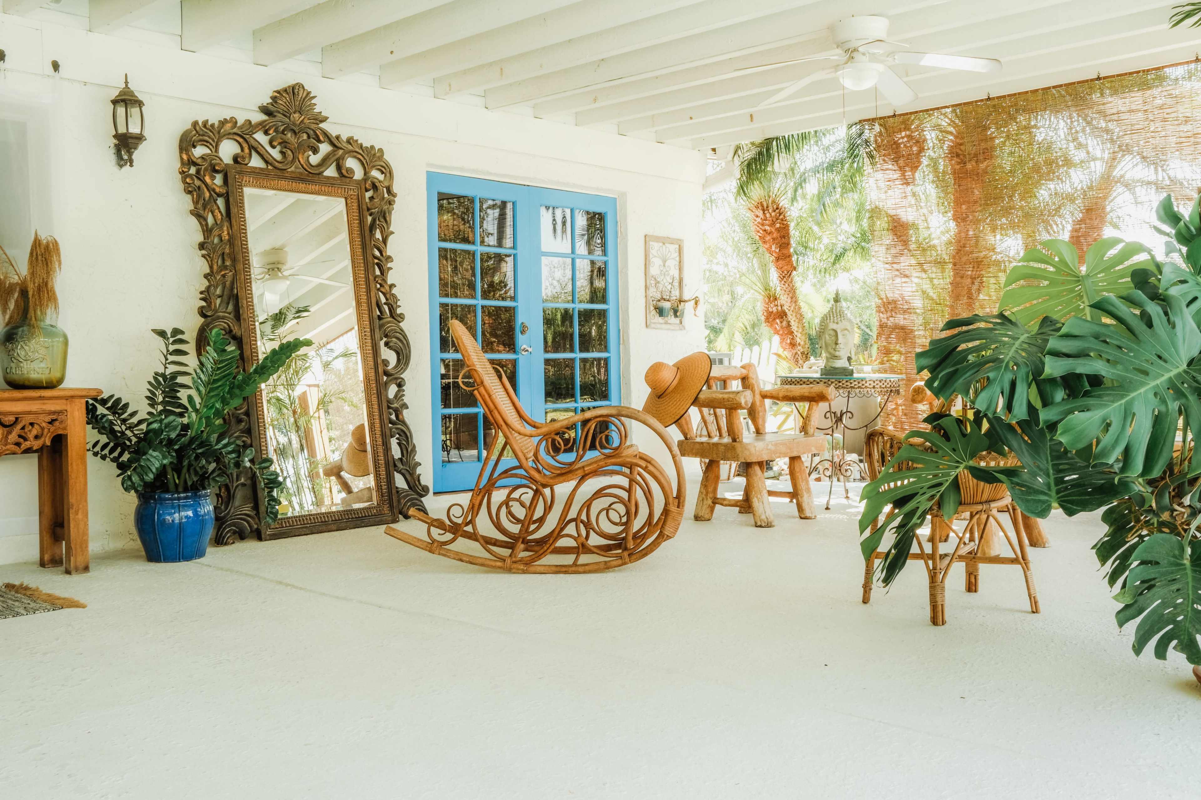 The image shows a covered patio with a wicker rocking chair, a large decorative mirror, a wooden table and chairs, and various plants.
