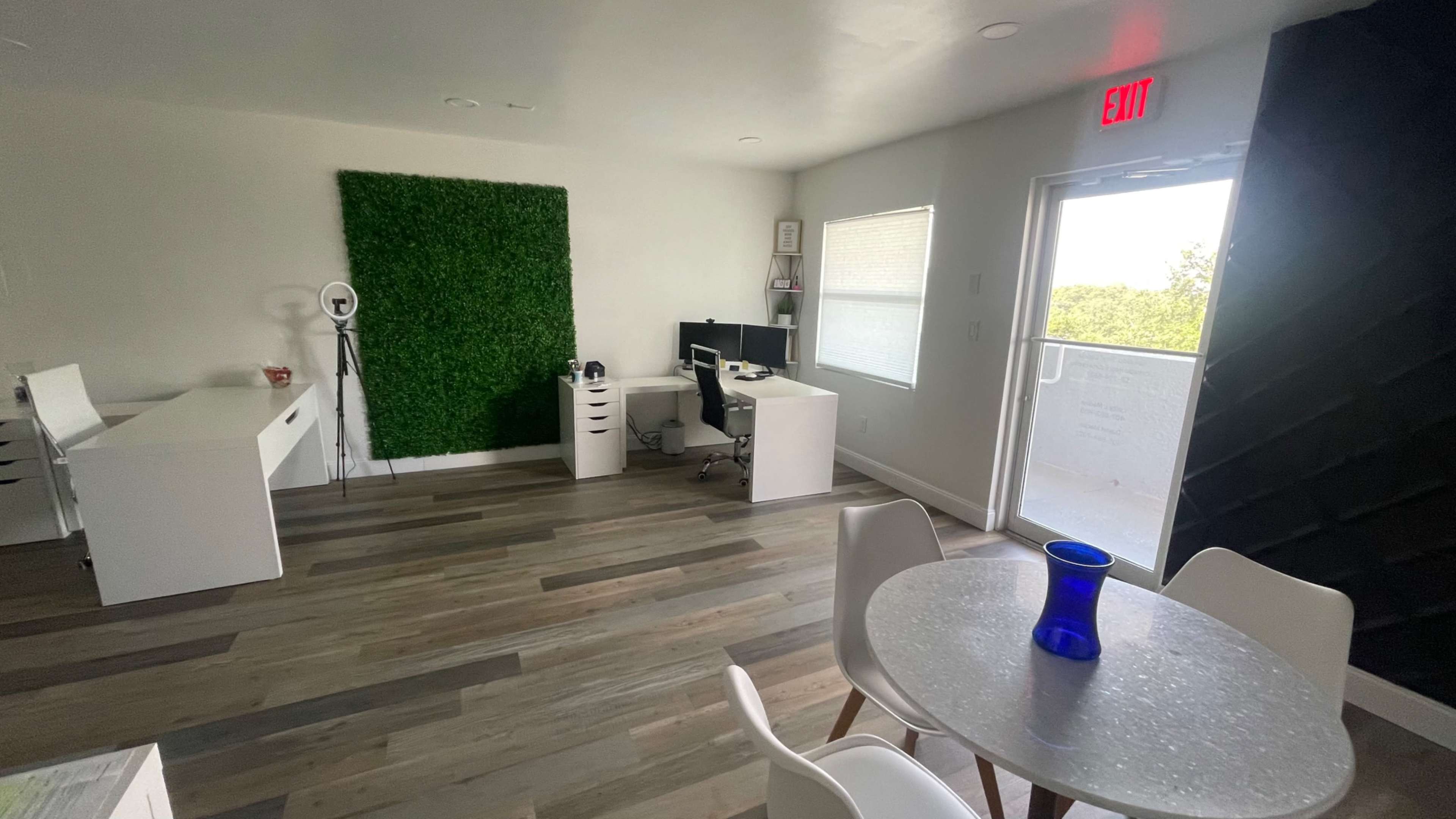 A modern office space featuring a desk with a computer, a circular dining table with a blue vase, and a wall covered in green foliage.