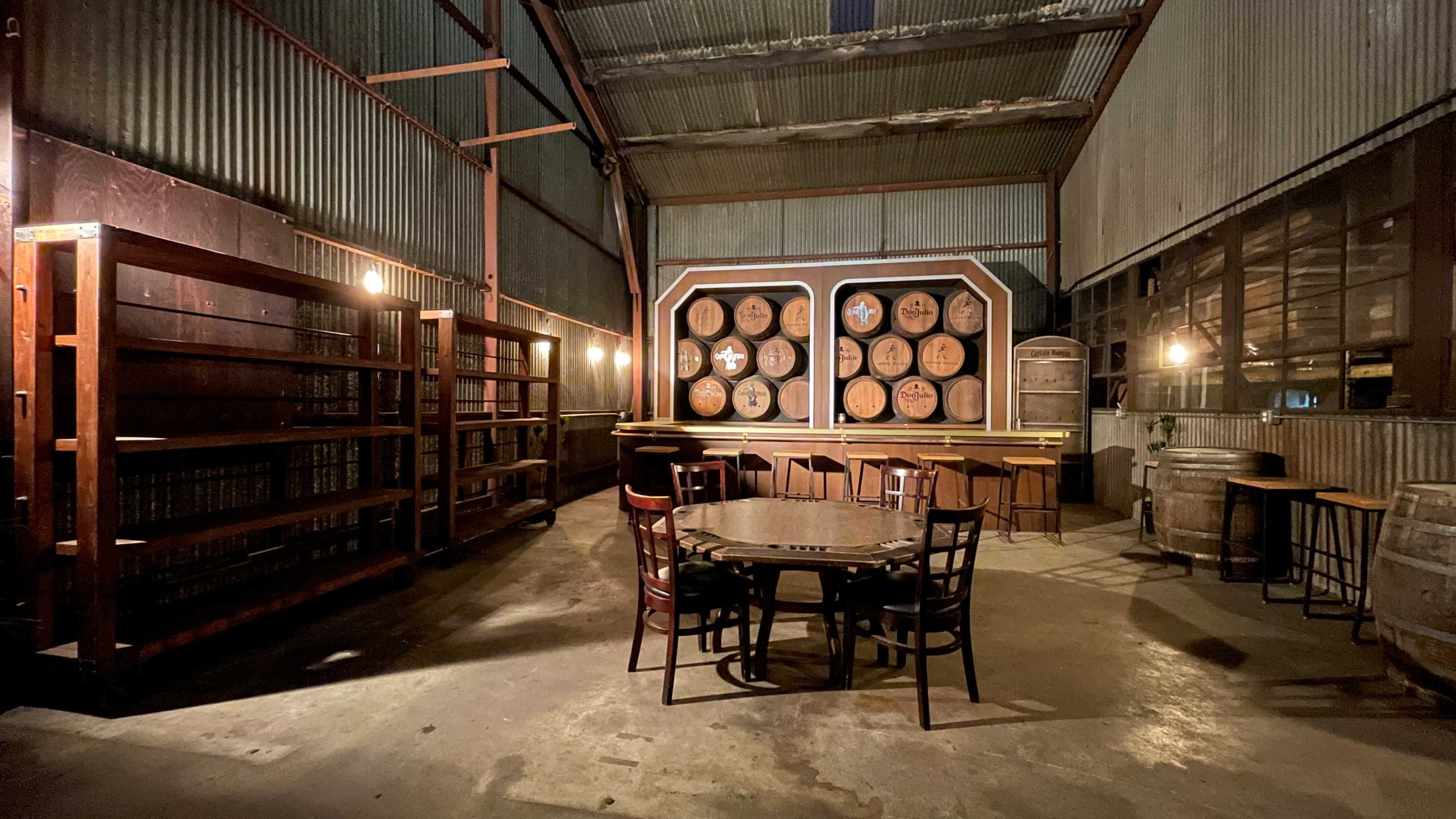 The interior of a dimly lit industrial warehouse features wooden shelves, a round table with chairs, and large barrels alongside a wall of stacked barrels.
