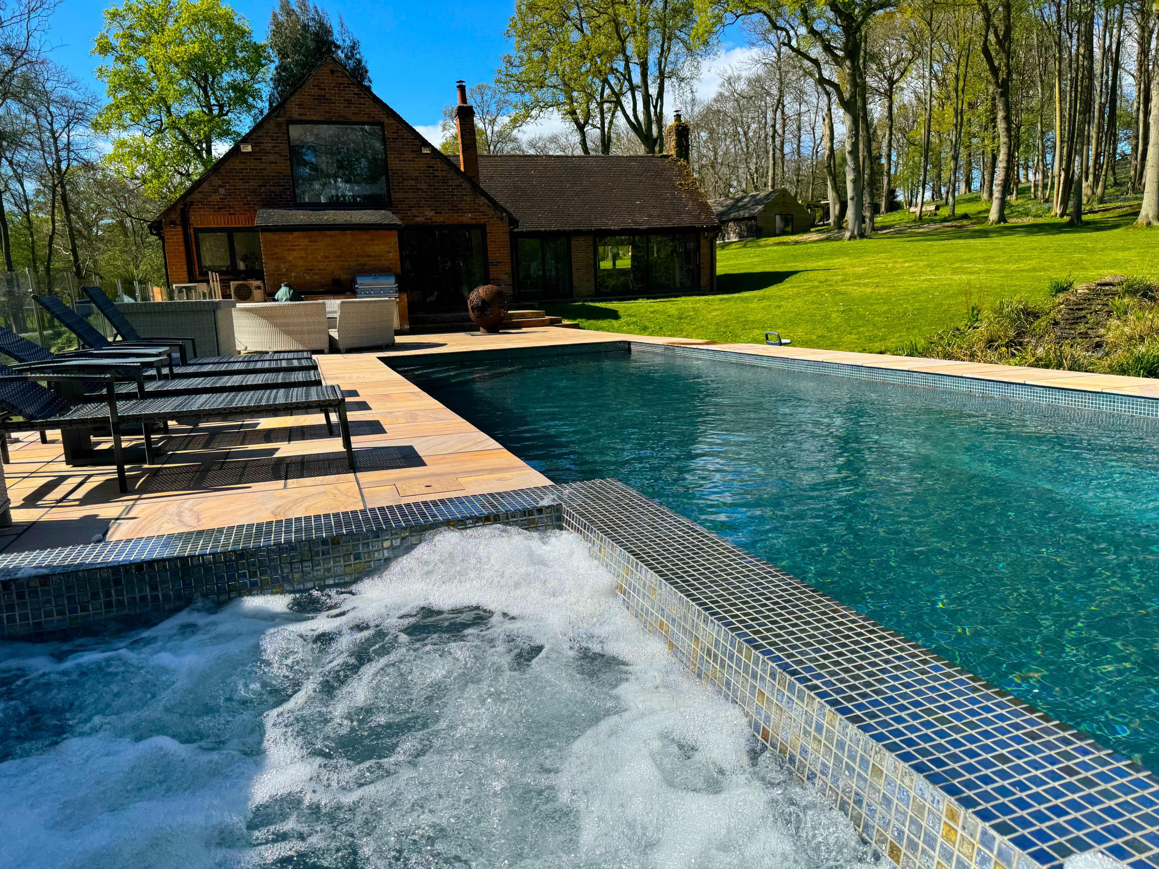 A clear swimming pool with a spa area is situated in front of a wooden house, surrounded by green trees and grass.