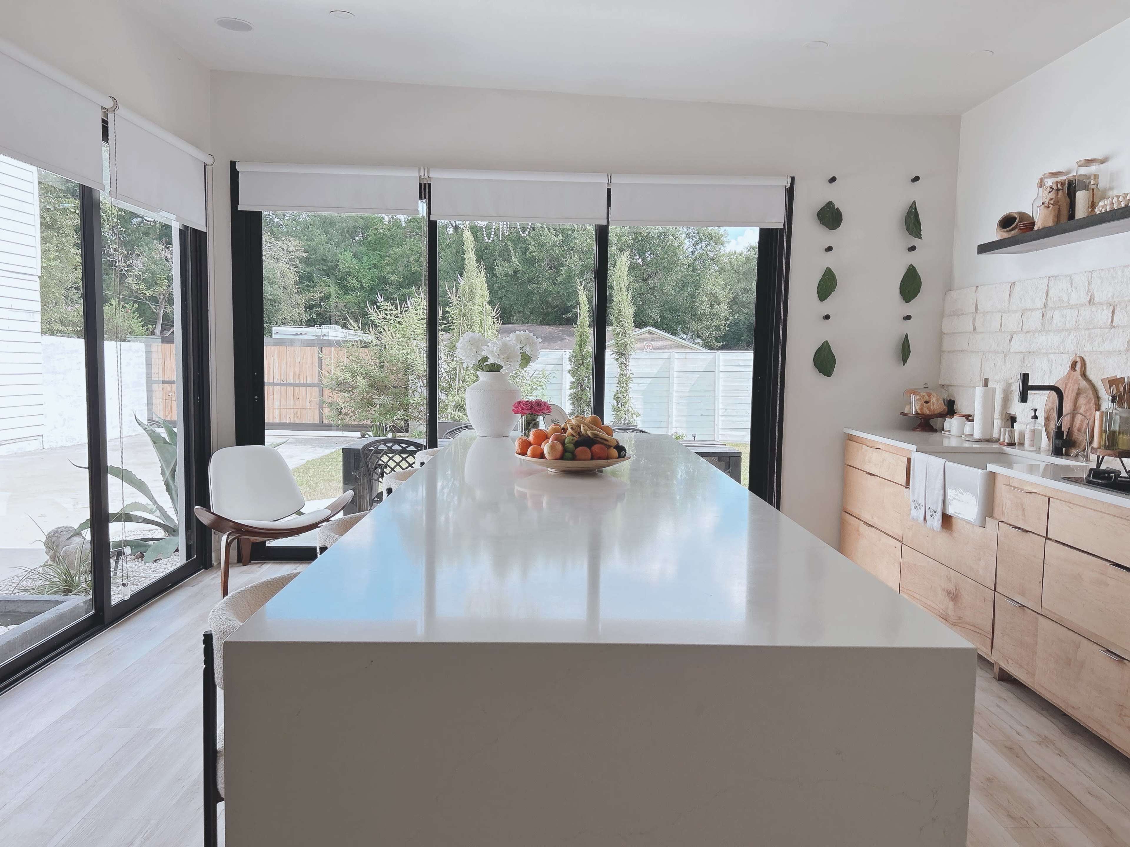 The image shows a modern kitchen with a large white countertop, wooden cabinetry, and large glass doors leading to an outdoor area.