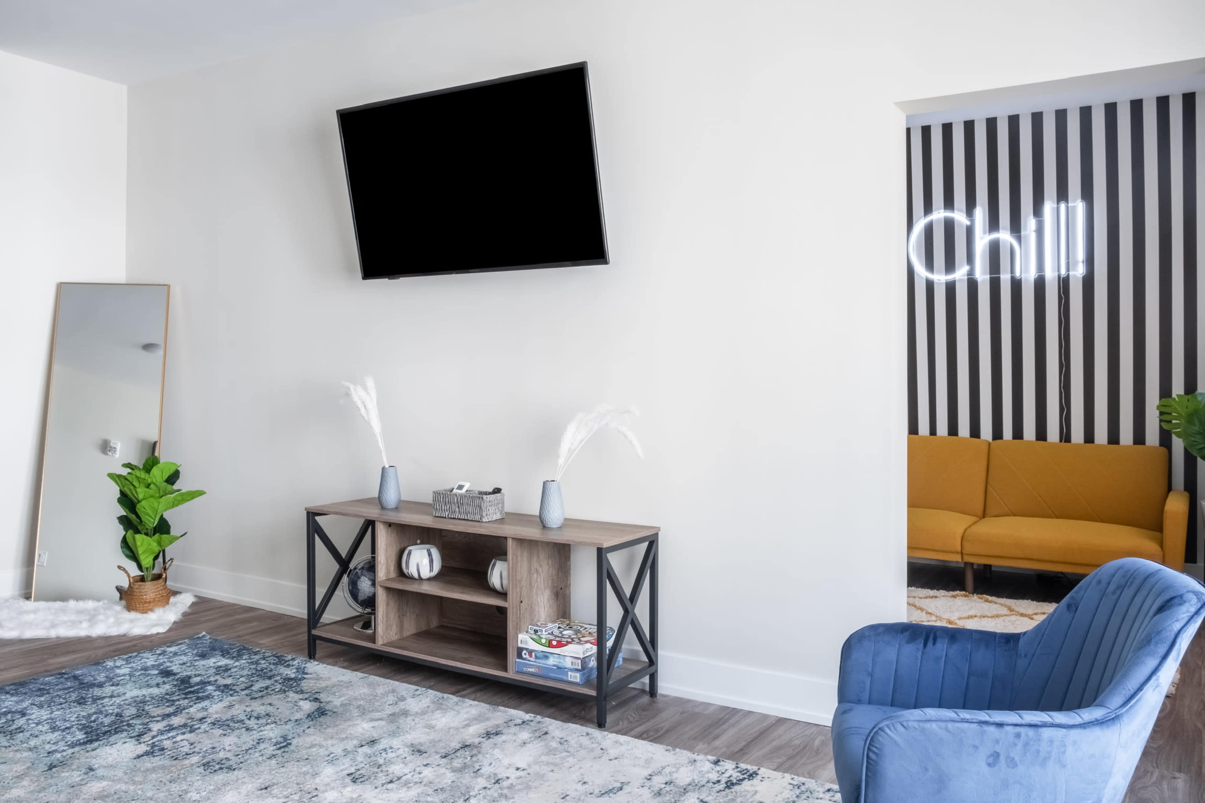 A modern living room features a television mounted on a wall, a wooden console table with decorative items, and a blue armchair beside a mirror, with a cozy seating area visible in an adjacent nook.