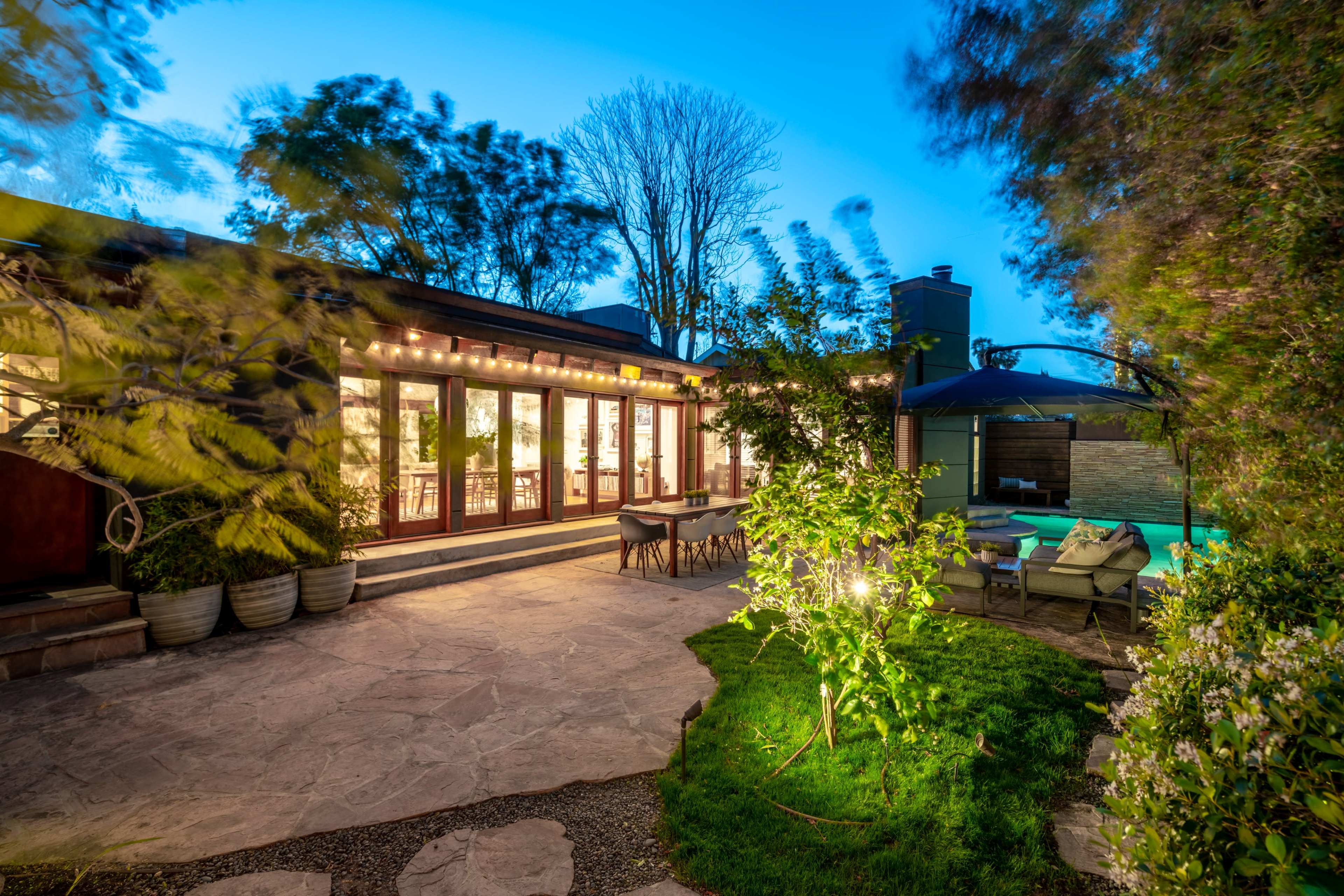 A modern home with large glass doors opens to a landscaped yard featuring trees, a seating area, and a swimming pool under an evening sky.