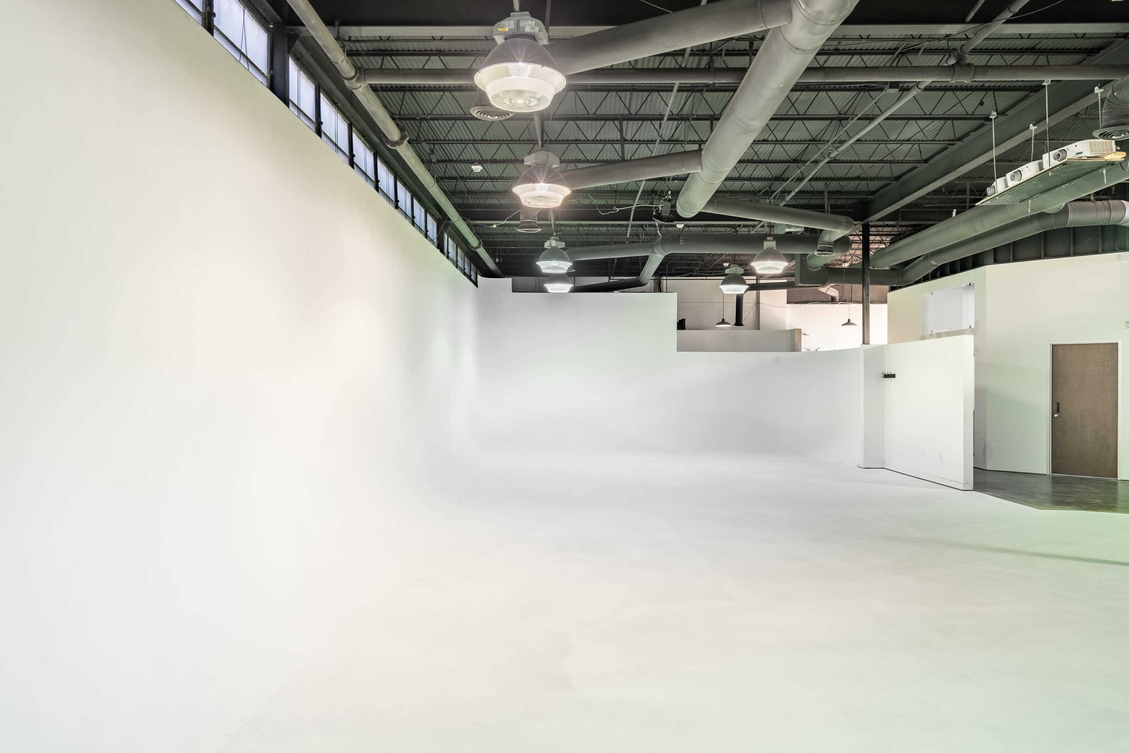 A large, empty studio space with white walls, a smooth floor, and industrial lighting fixtures.