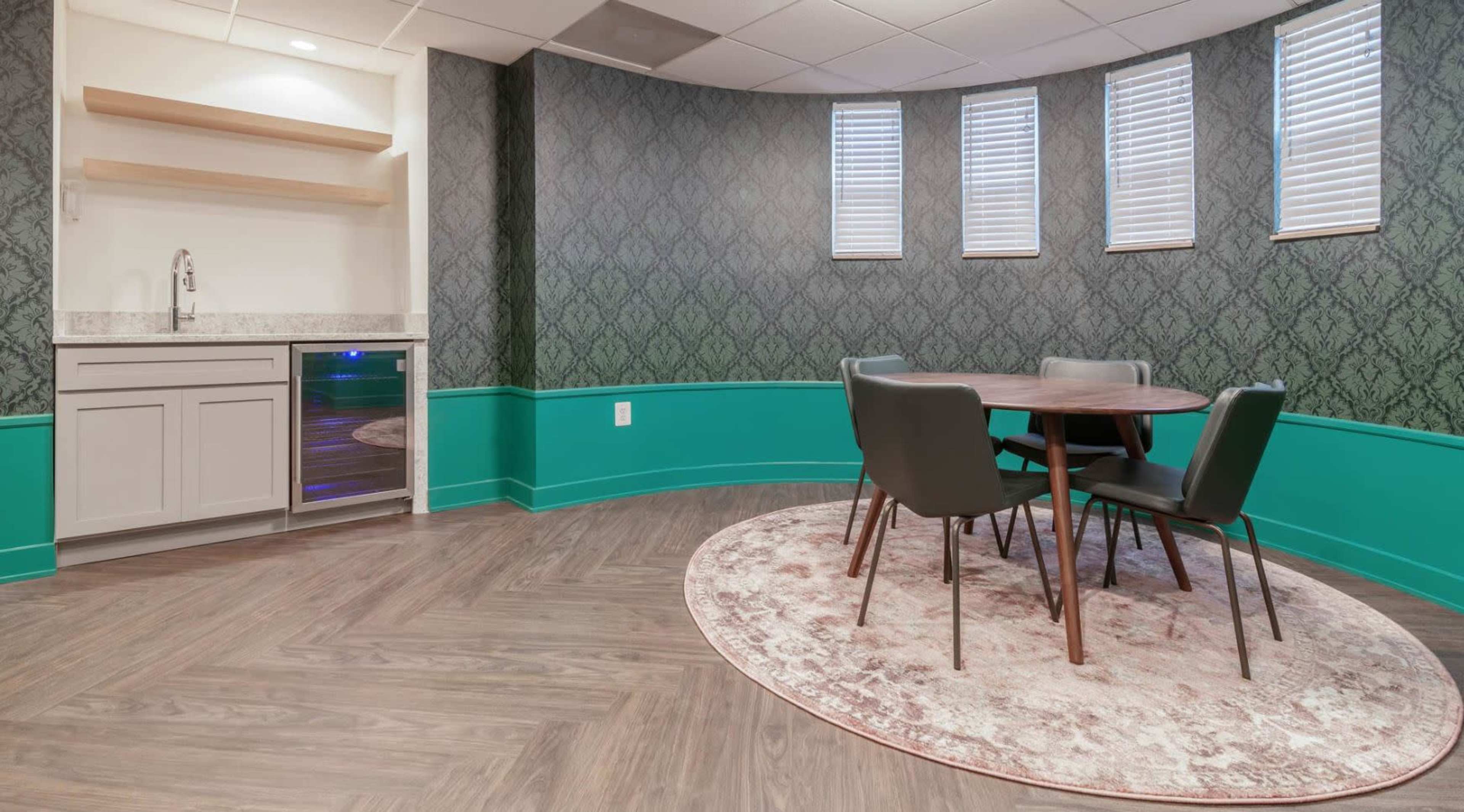 A small dining area with a round wooden table and four dark chairs, surrounded by patterned wallpaper and featuring a kitchenette with a sink and glass door refrigerator.