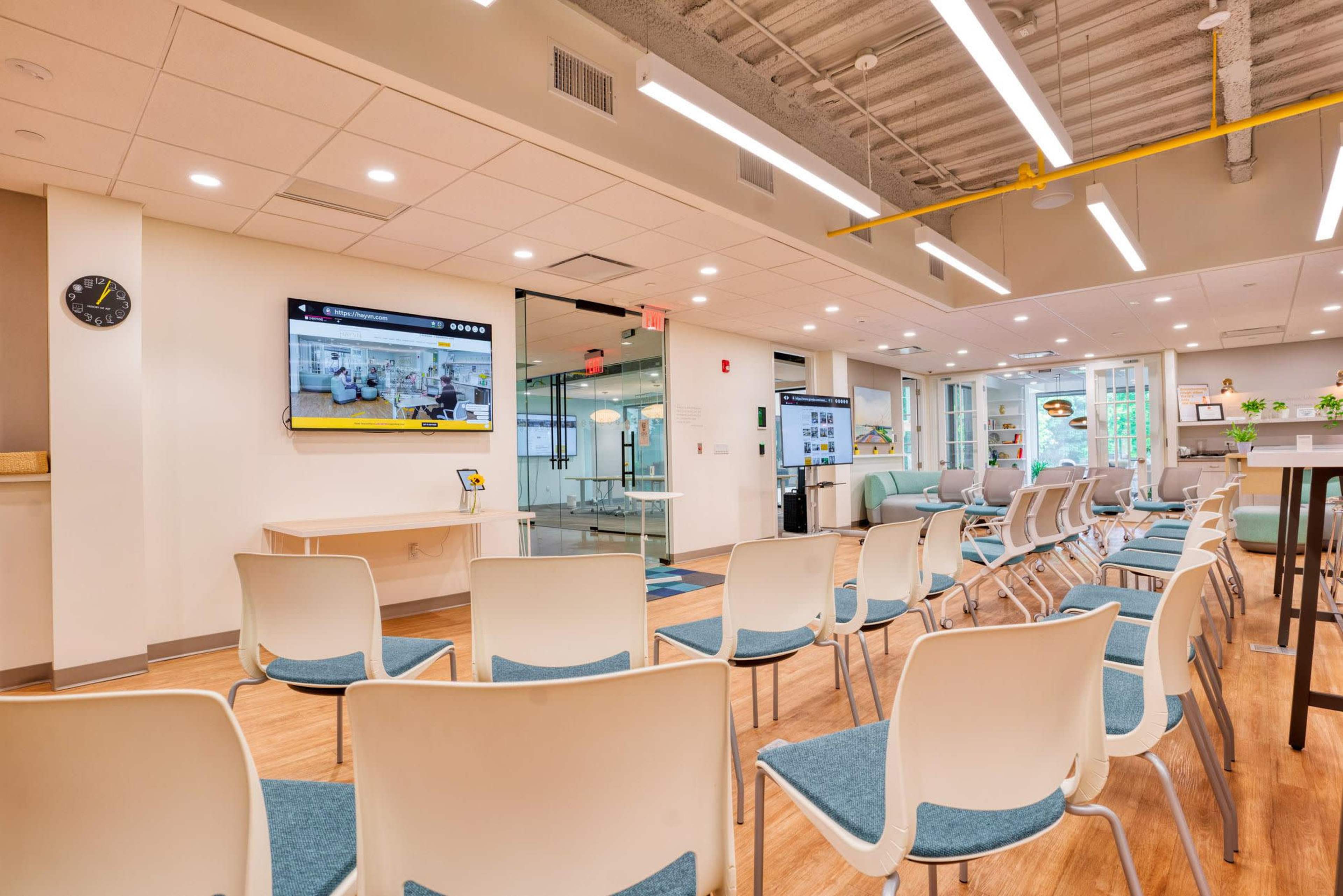A waiting room with white chairs arranged in rows, a wall-mounted television displaying content, and a clock on the wall.