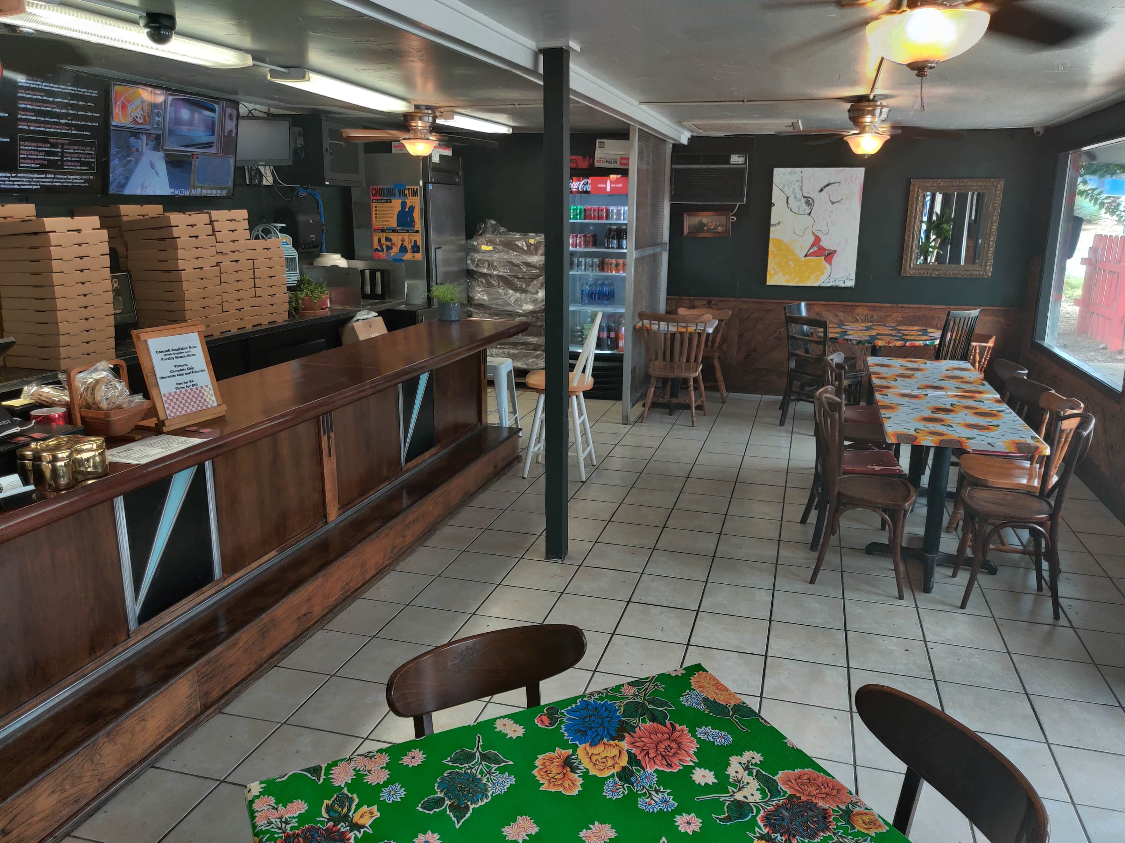 A small, cozy restaurant interior with wooden tables, a counter filled with pizza boxes in the background, and colorful tablecloths.