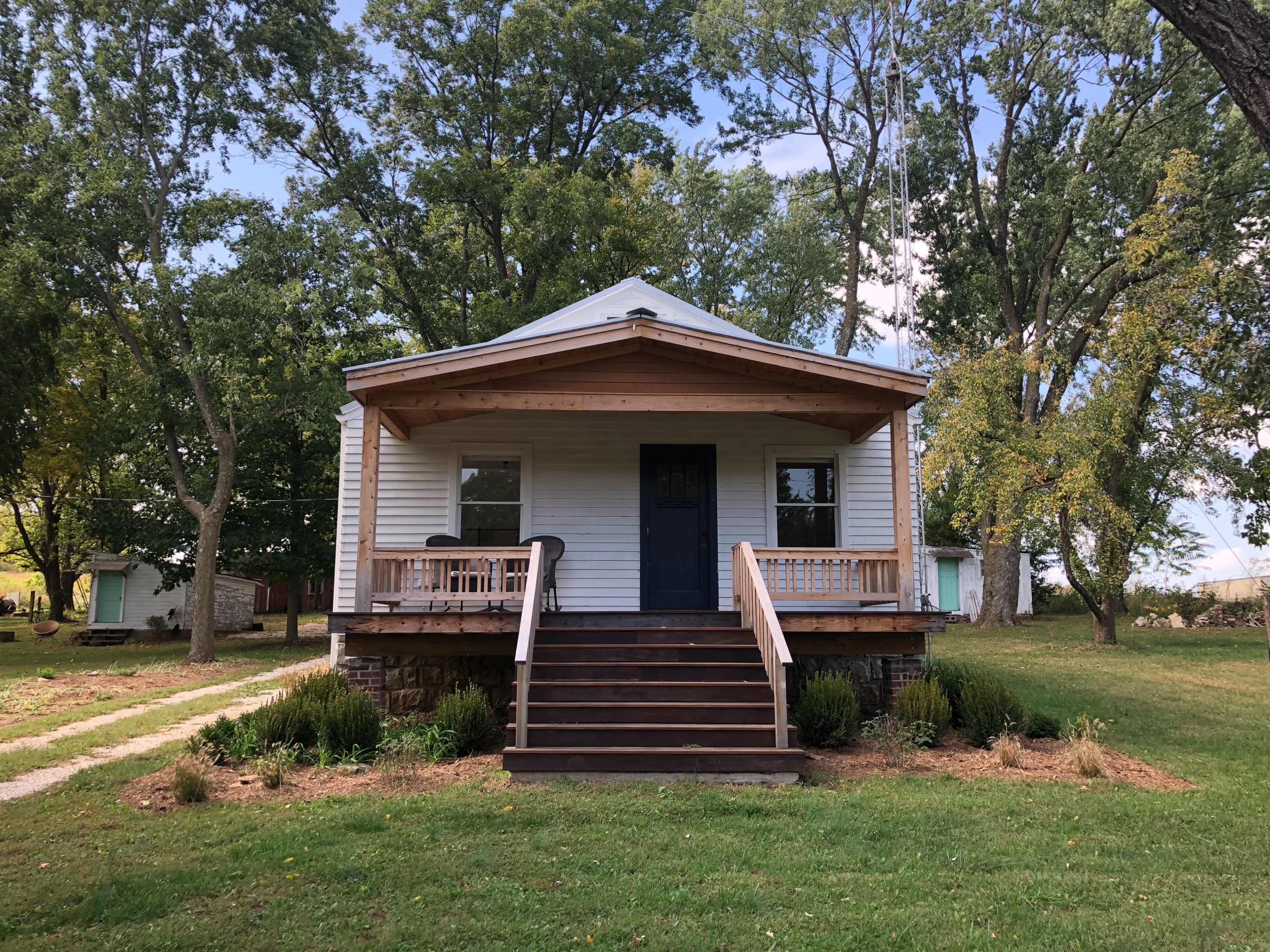 A small white house with a front porch and wooden steps is surrounded by green grass and trees.