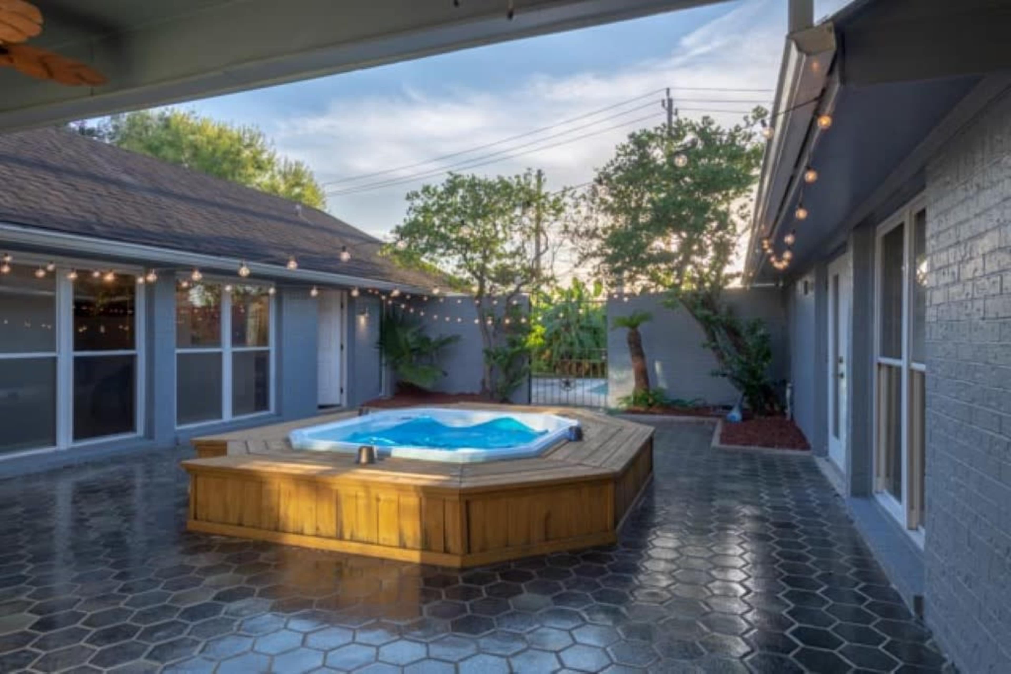 The image shows a courtyard featuring a wooden hot tub surrounded by hexagonal tiled flooring and string lights, with lush greenery visible in the background.