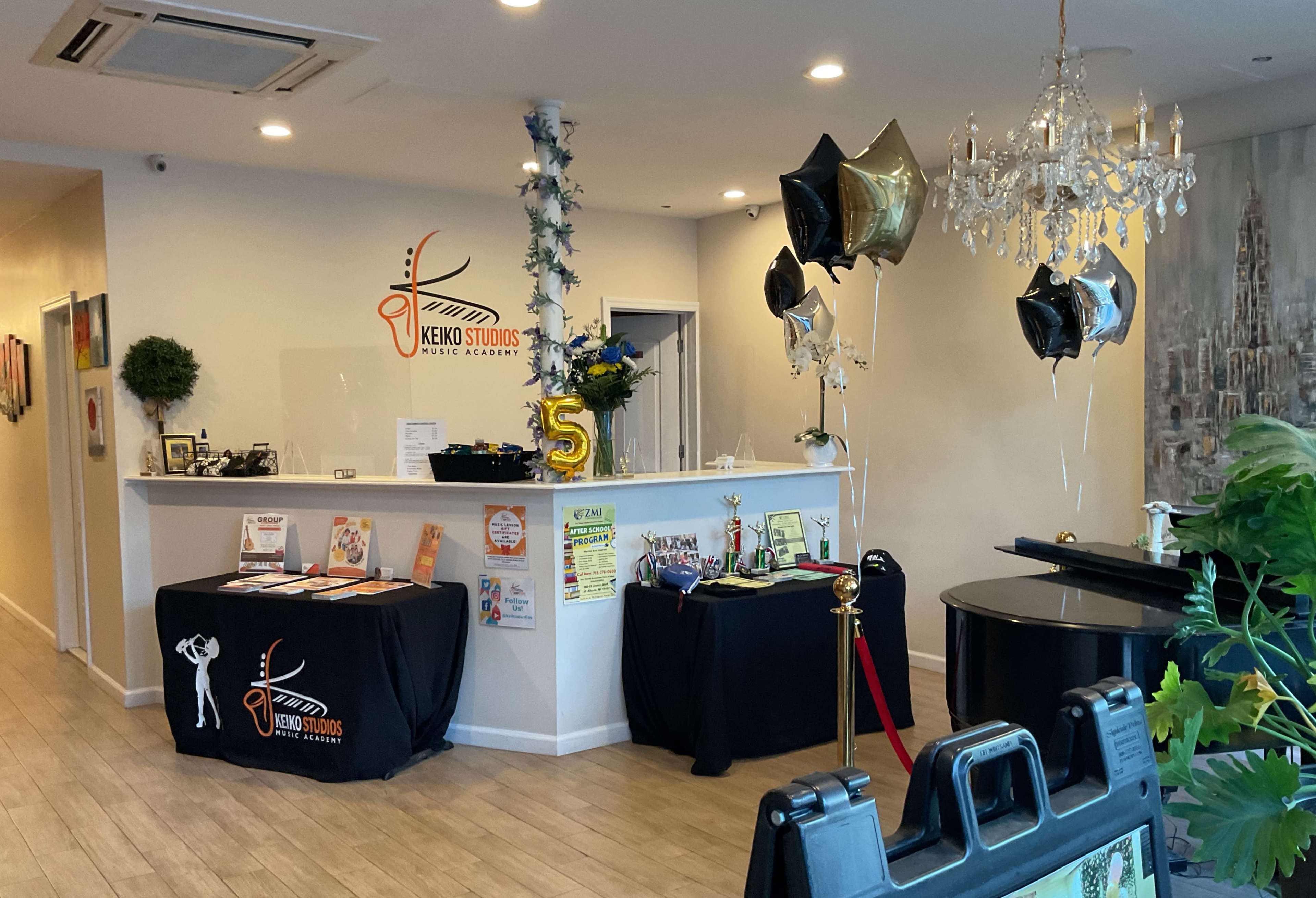 The image shows the interior of a music academy with a reception desk, black balloons, and a grand piano.