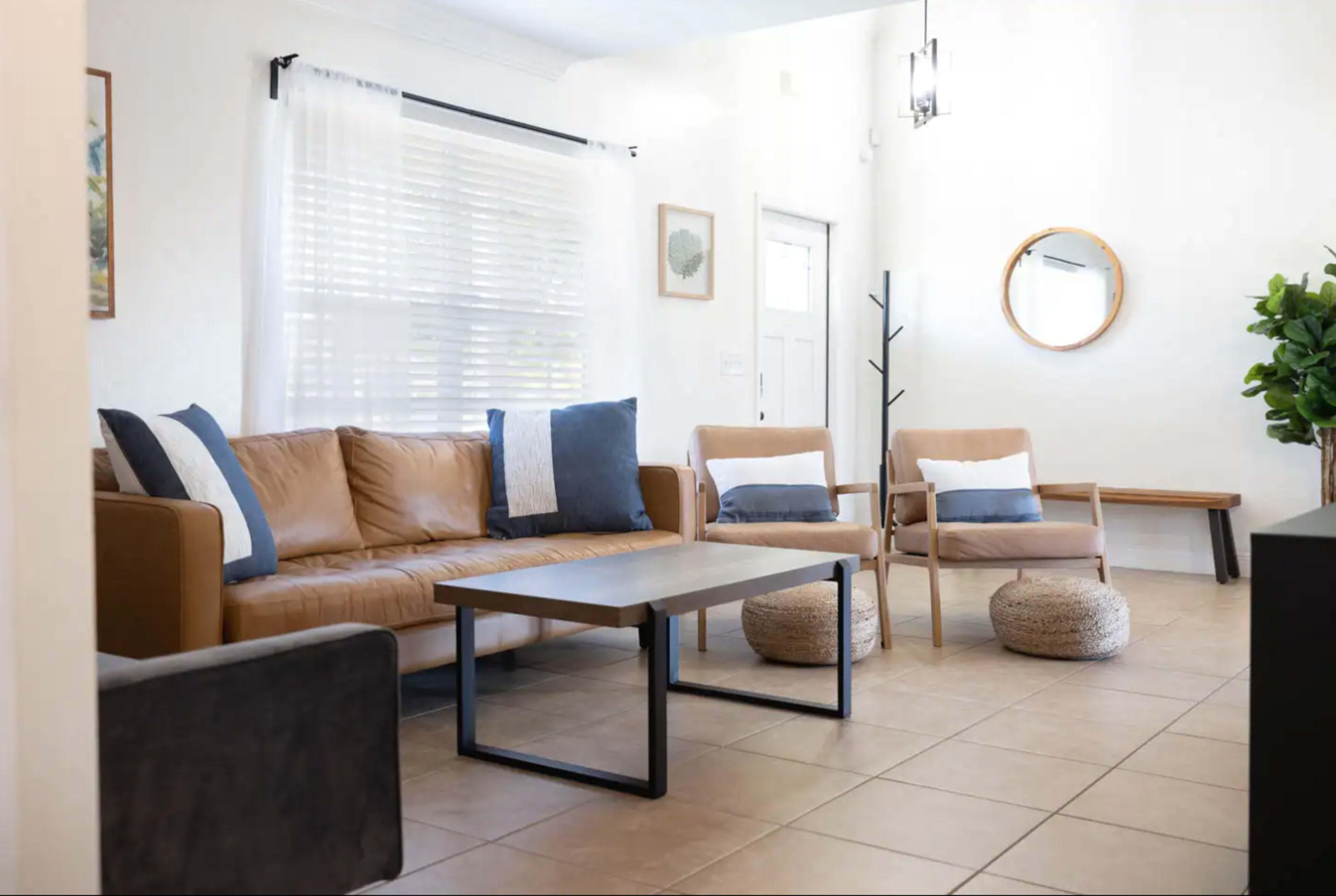 A living room features a brown leather sofa, two armchairs, a round mirror, and a coffee table on tiled flooring.