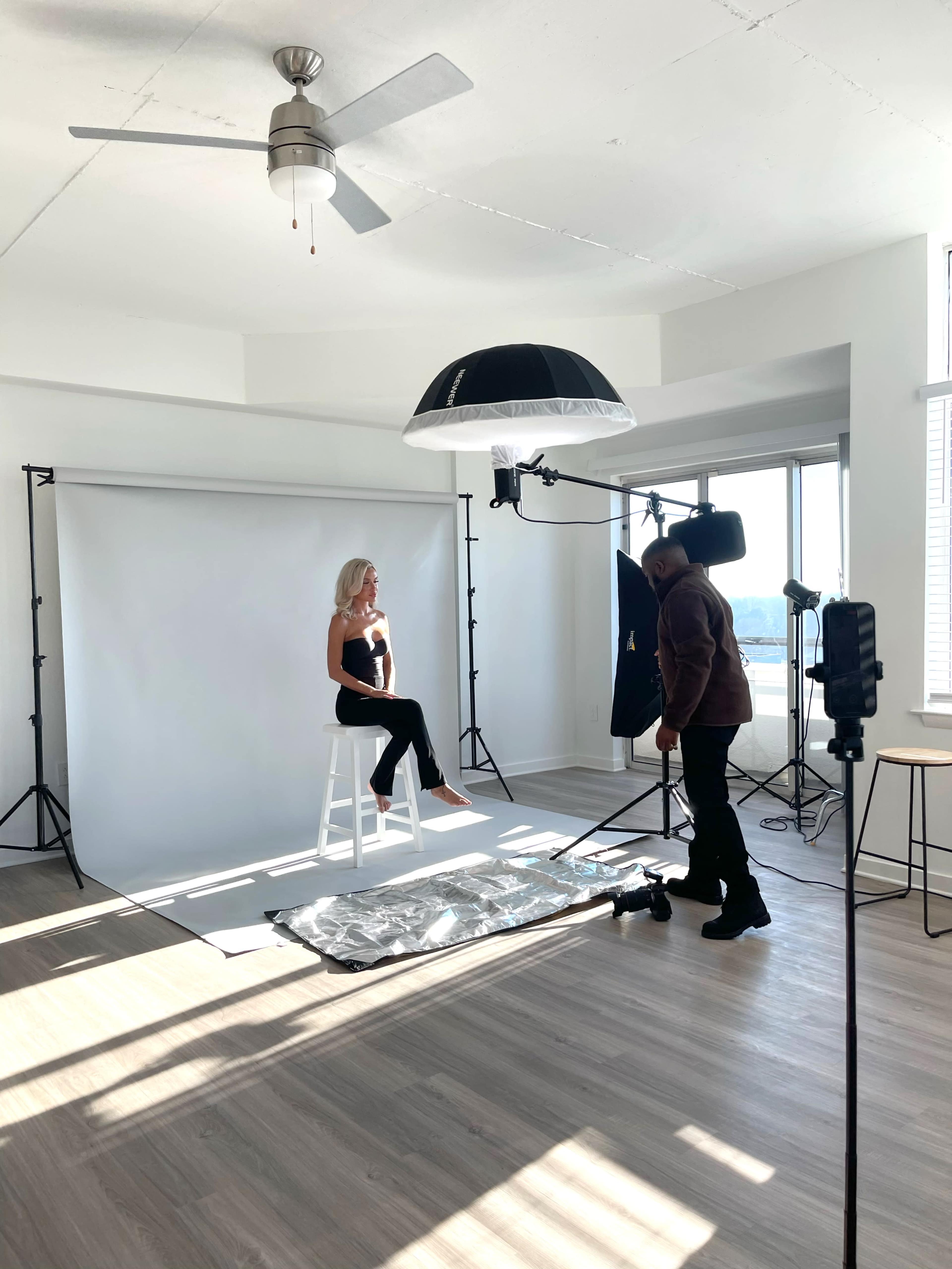 A model sits on a white stool in front of a backdrop while a photographer adjusts lighting equipment in a well-lit studio space.
