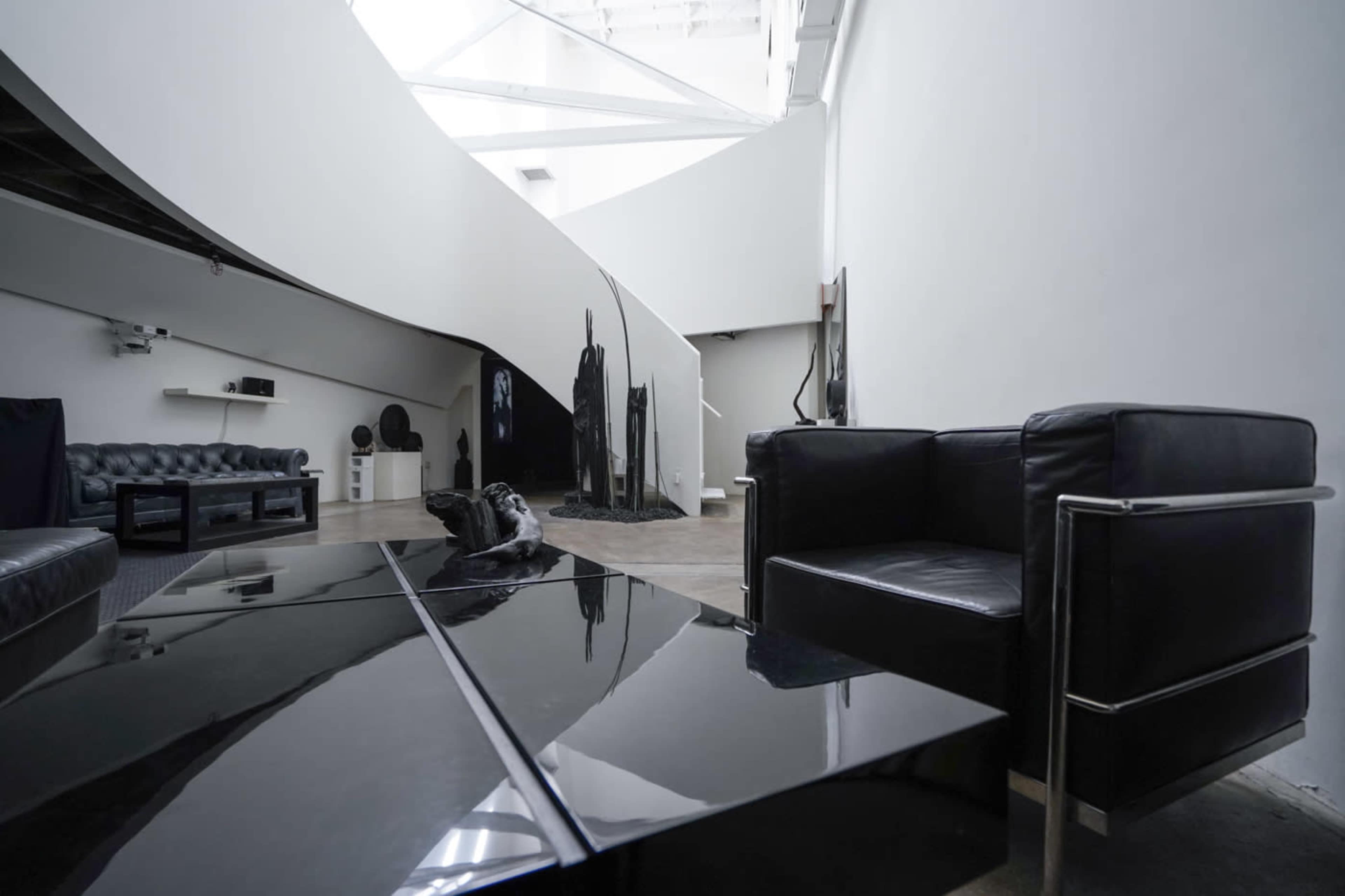 The image shows a modern, minimalist interior featuring a black leather sofa and armchair, a sleek black coffee table, and a curved white wall extending into a large, open space.