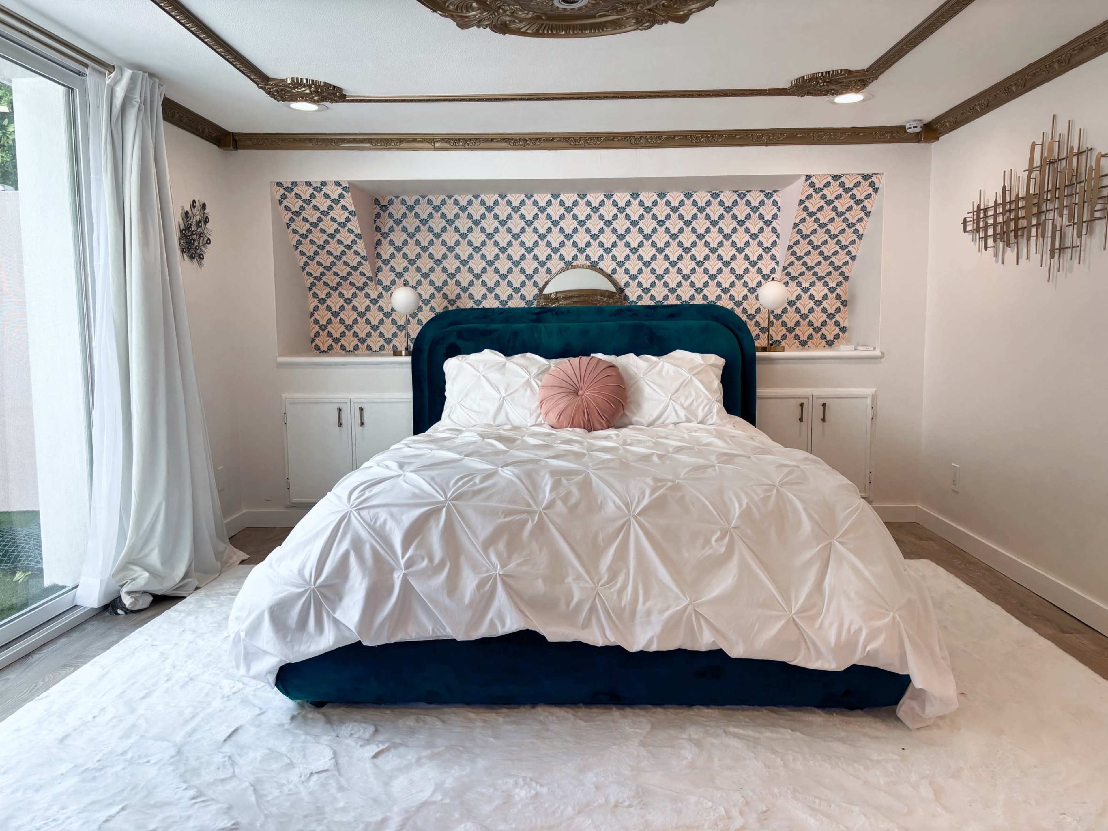 The image shows a modern bedroom with a blue velvet bed, white bedding, and patterned wallpaper behind the headboard.