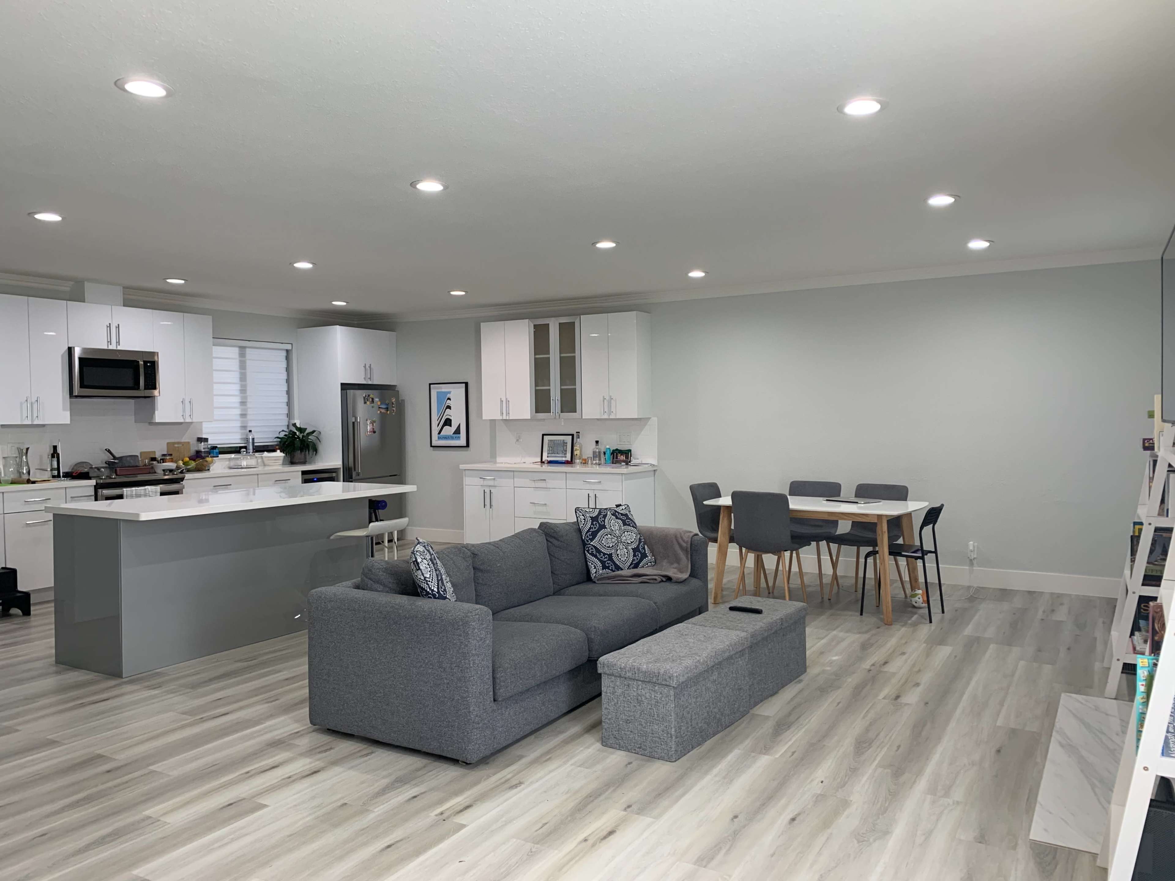 A modern, open-concept living space featuring a gray sectional sofa, a dining table, and a kitchen with white cabinetry and appliances.