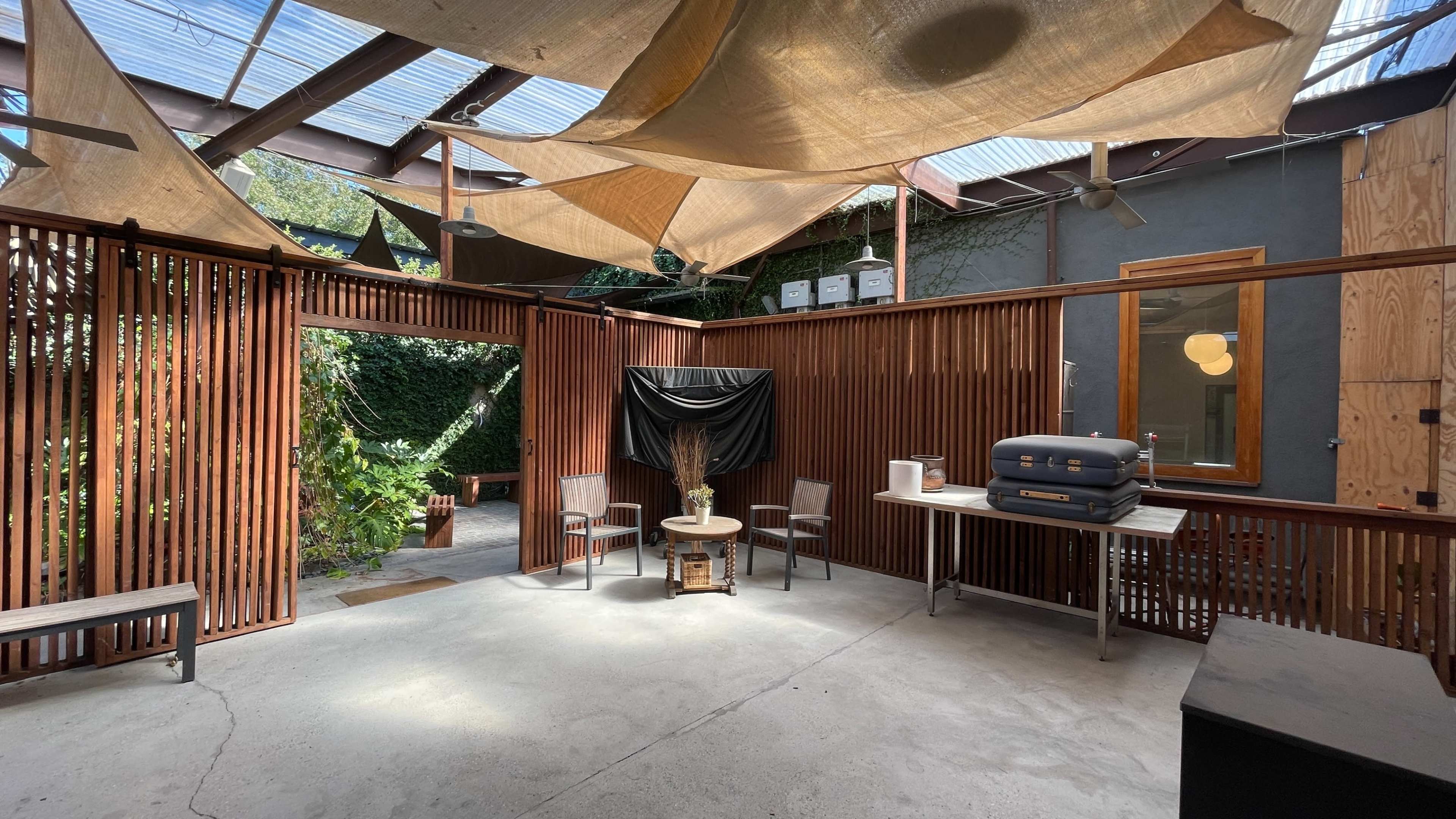 The image shows a modern, open indoor space with wooden slatted walls, two chairs, a small table, and lush greenery visible through an opening leading to an outdoor area.