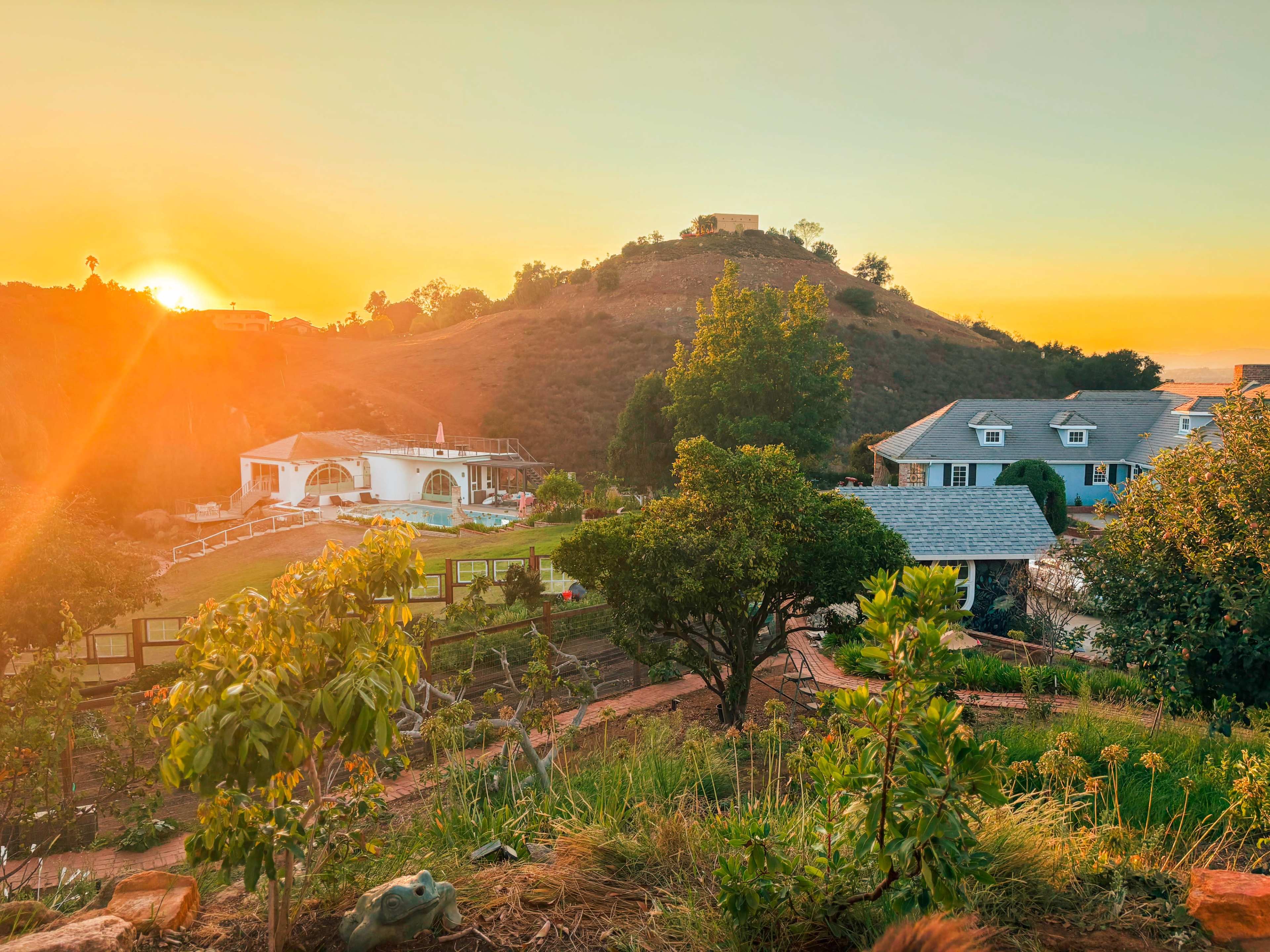 The image shows a scenic view of a hillside at sunset, with houses and gardens in the foreground and a bright orange sky in the background.