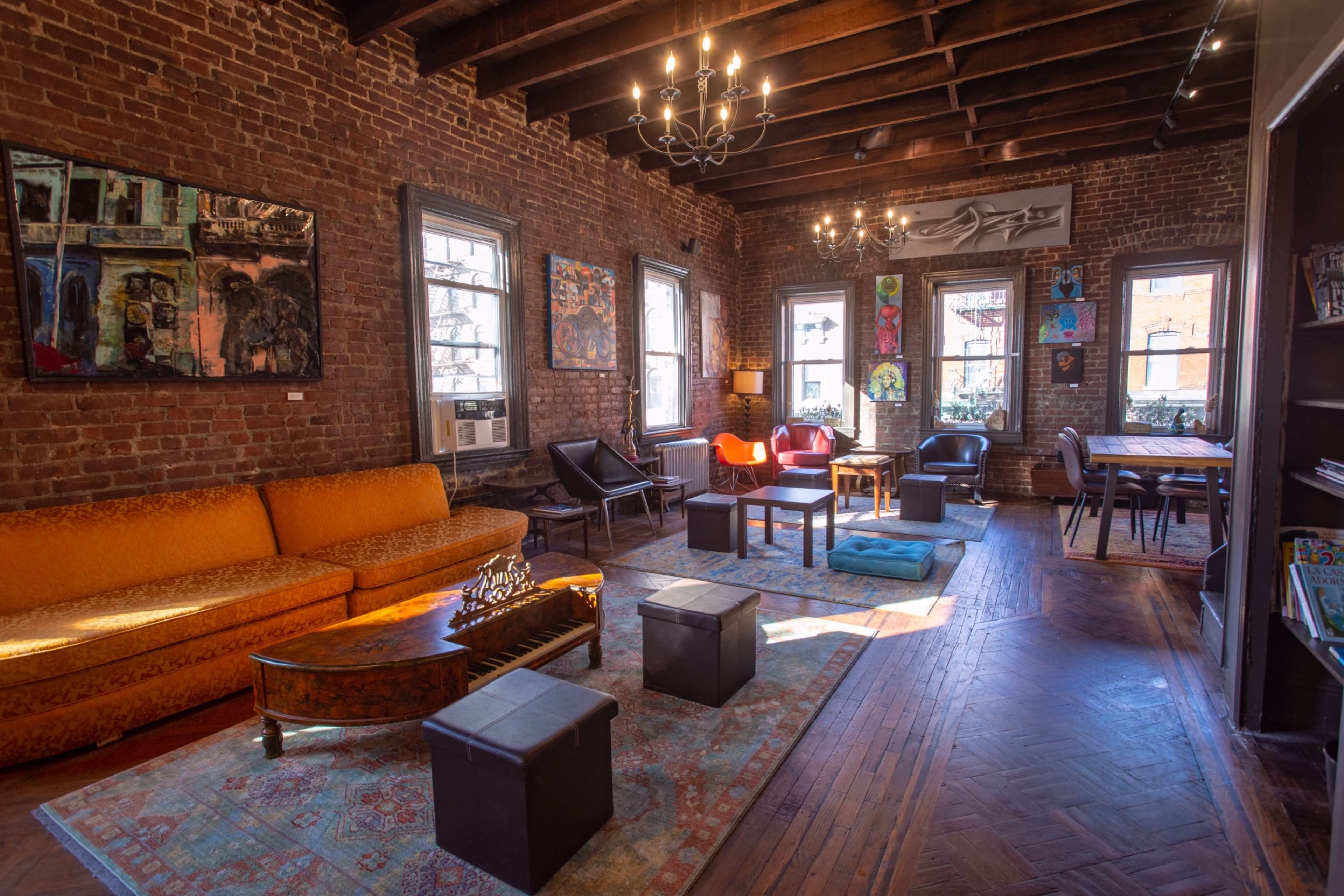 The room features exposed brick walls, a large orange sofa, various artworks, and a mix of seating arrangements, illuminated by overhead lighting.