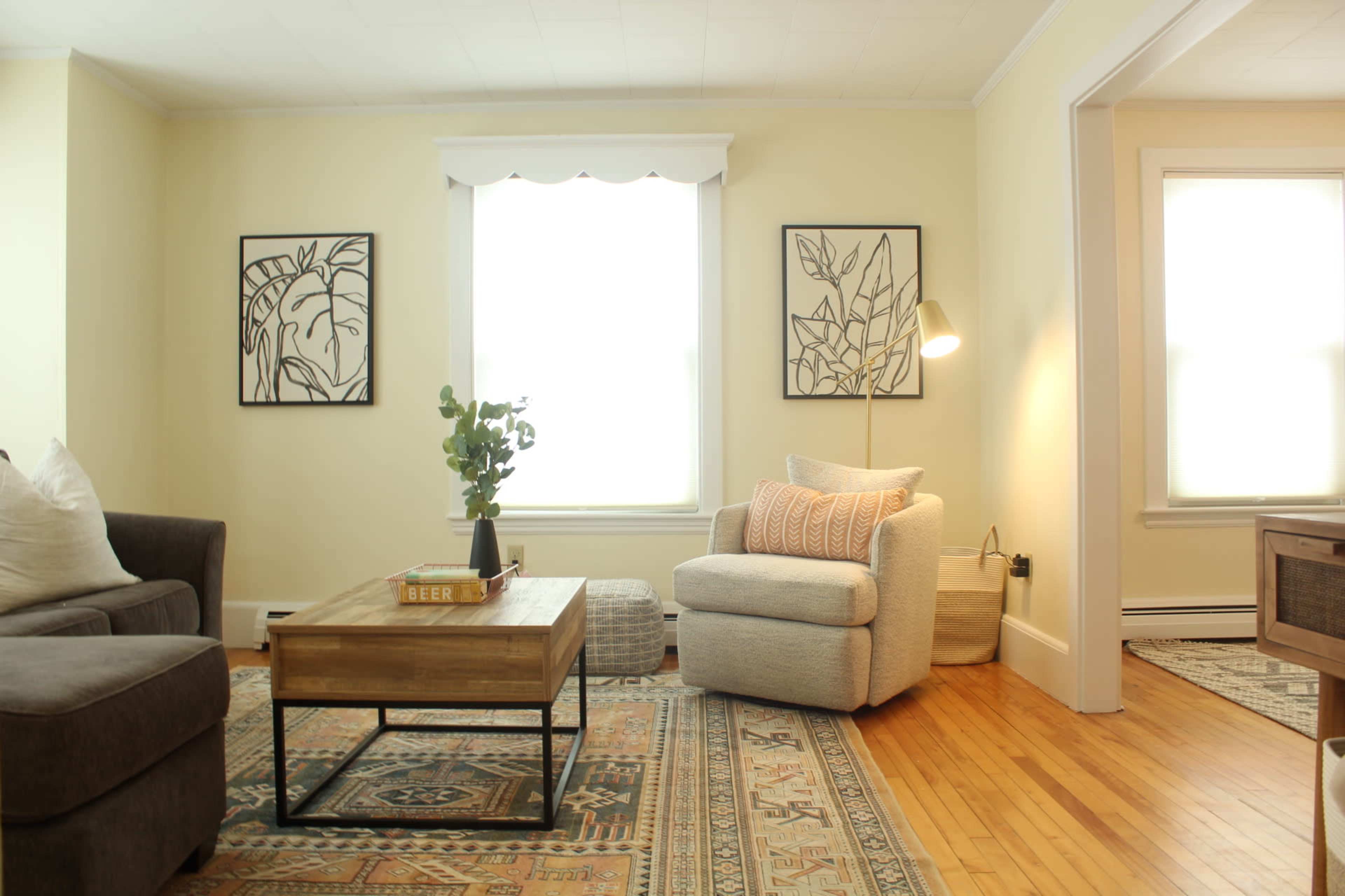 A modern living room features a gray sofa, a light-colored armchair, a wooden coffee table, and artwork on the walls, with large windows allowing natural light to fill the space.
