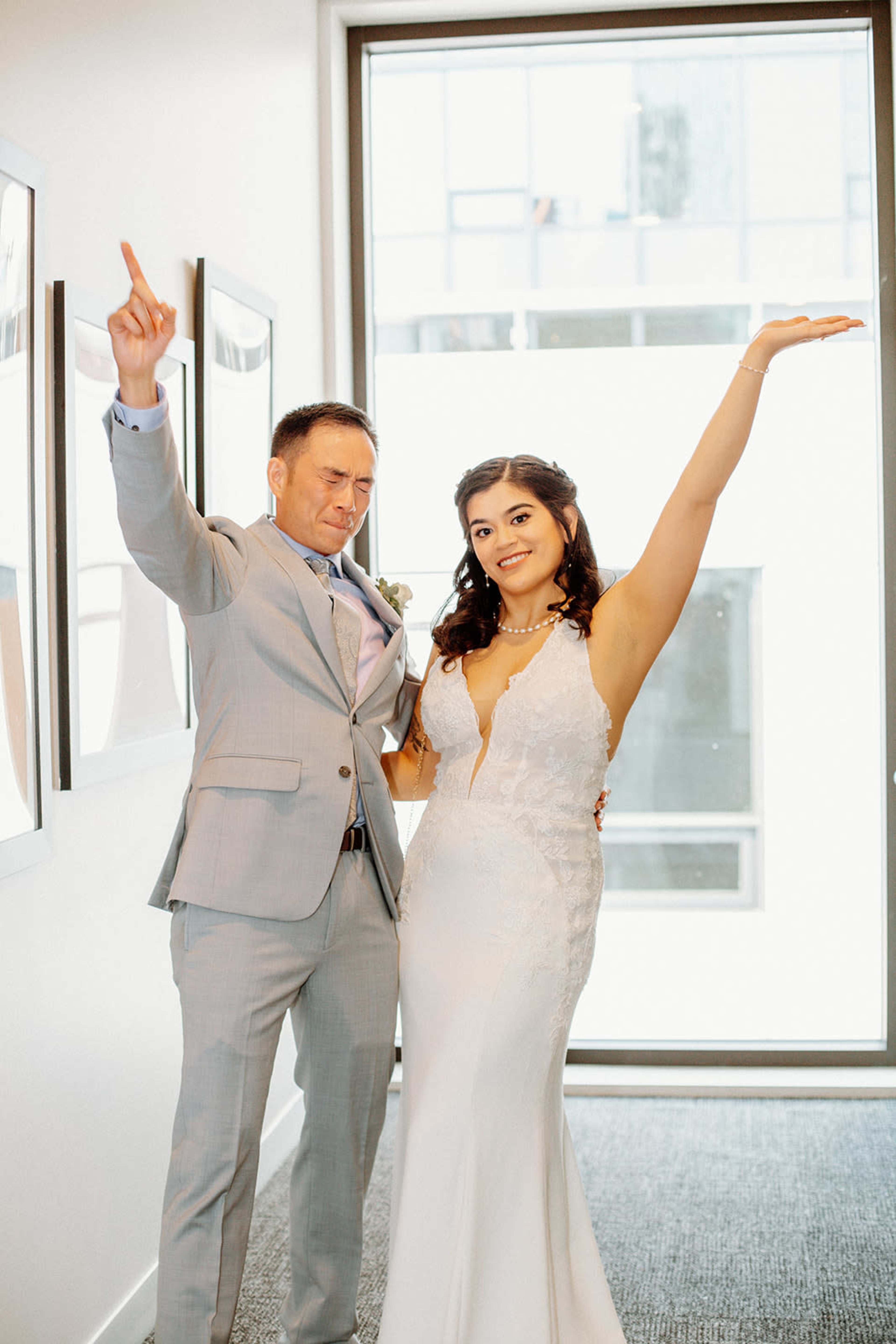 A man and woman pose together in formal attire, celebrating joyfully in a well-lit hallway with large windows.