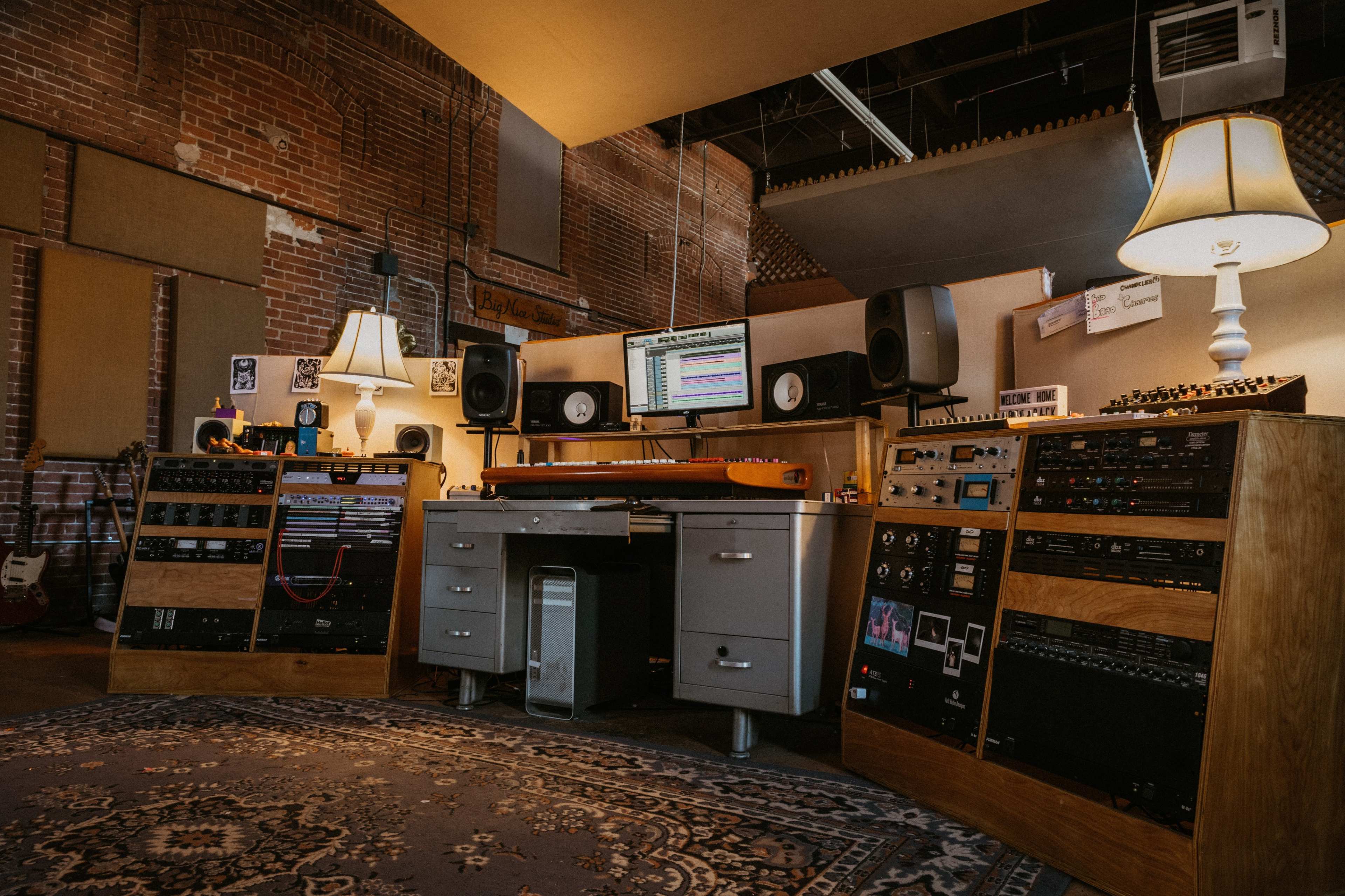 A recording studio with audio equipment, mixing consoles, a computer monitor displaying software, and ambient lighting from two lamps.