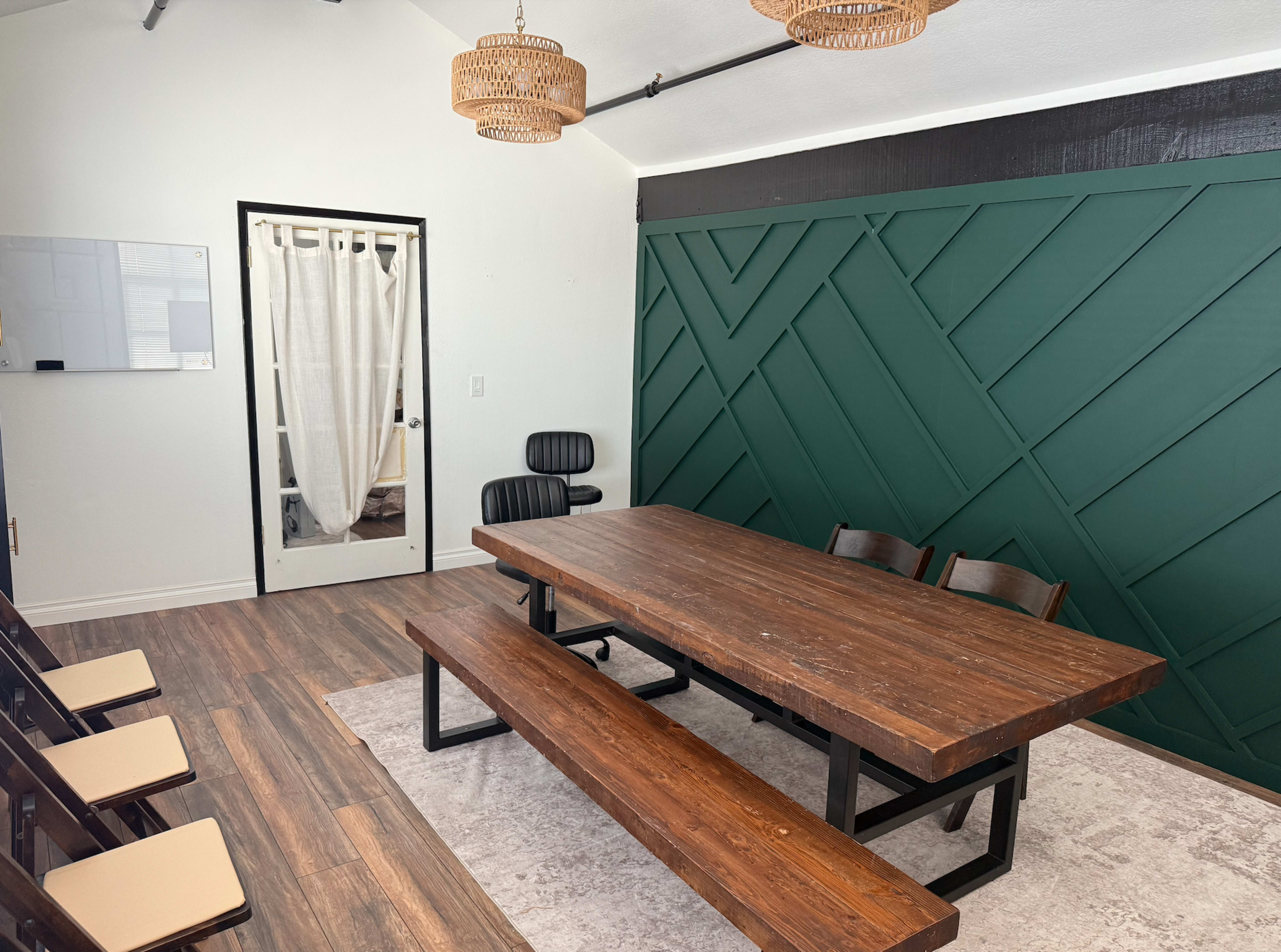 The image shows a meeting room featuring a large wooden table with benches, several chairs, a green accent wall, and a door with a curtain.