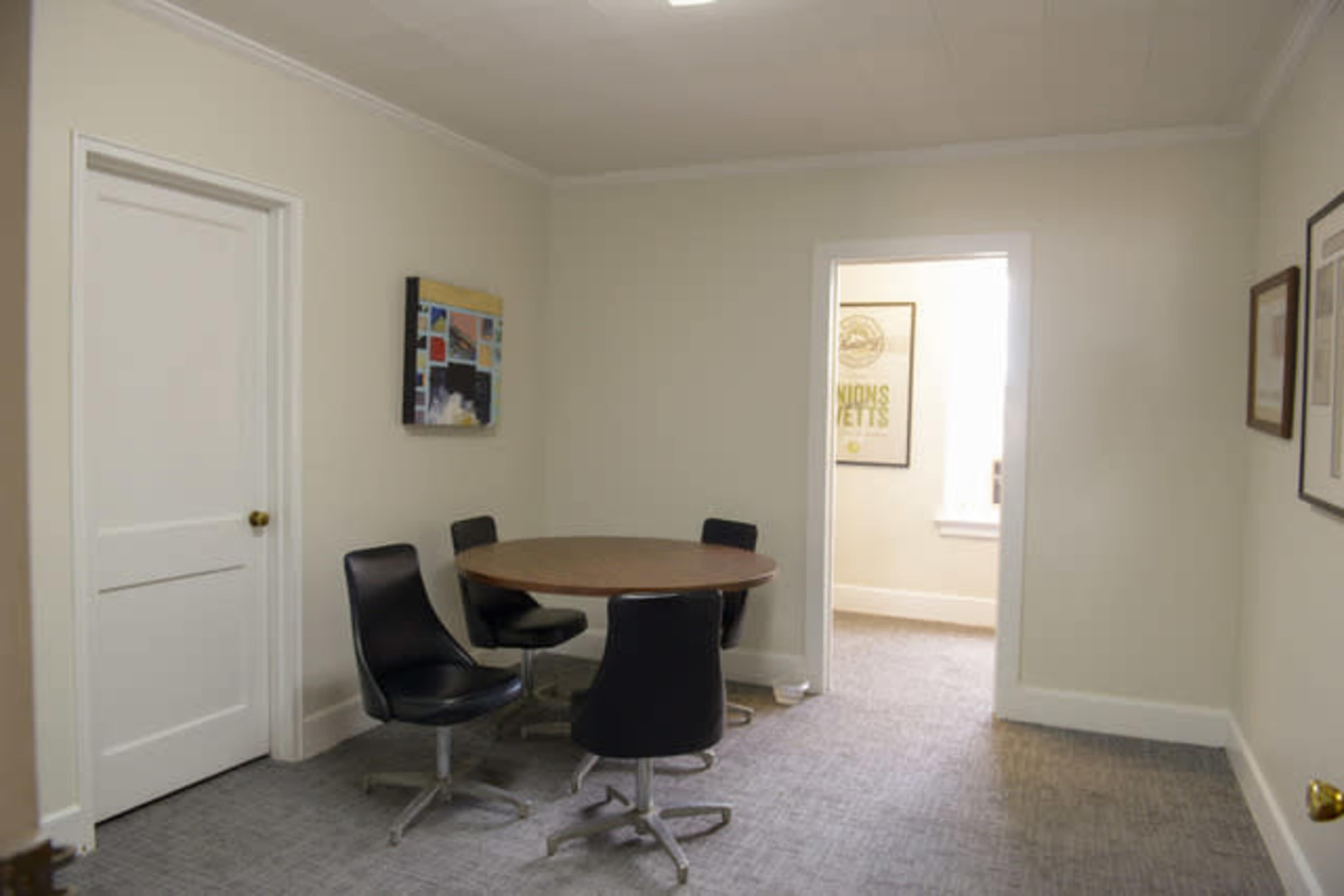 A small meeting room features a round table surrounded by four black chairs, with a door and doorway leading to another room.