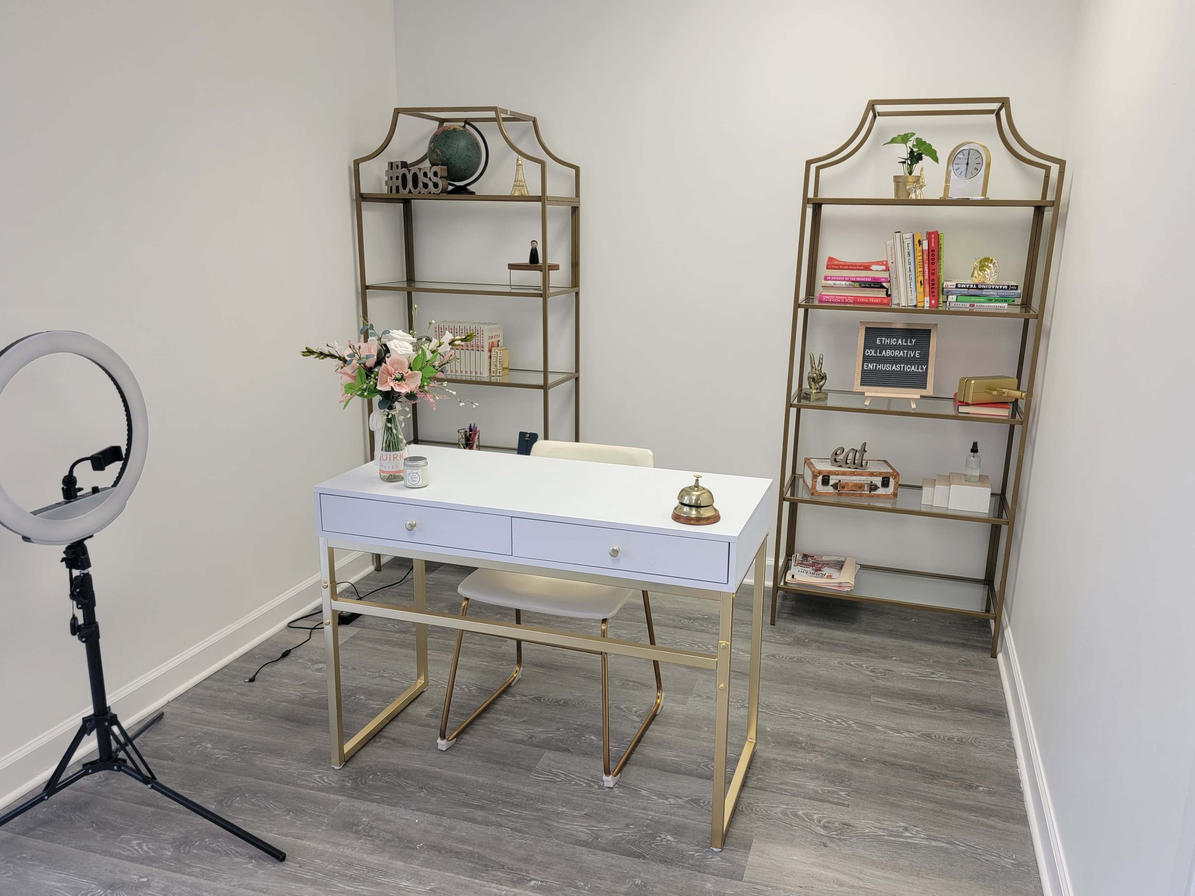 A modern office space featuring a white desk with gold accents, two open bookshelves filled with various items, and a ring light leaning against the wall.
