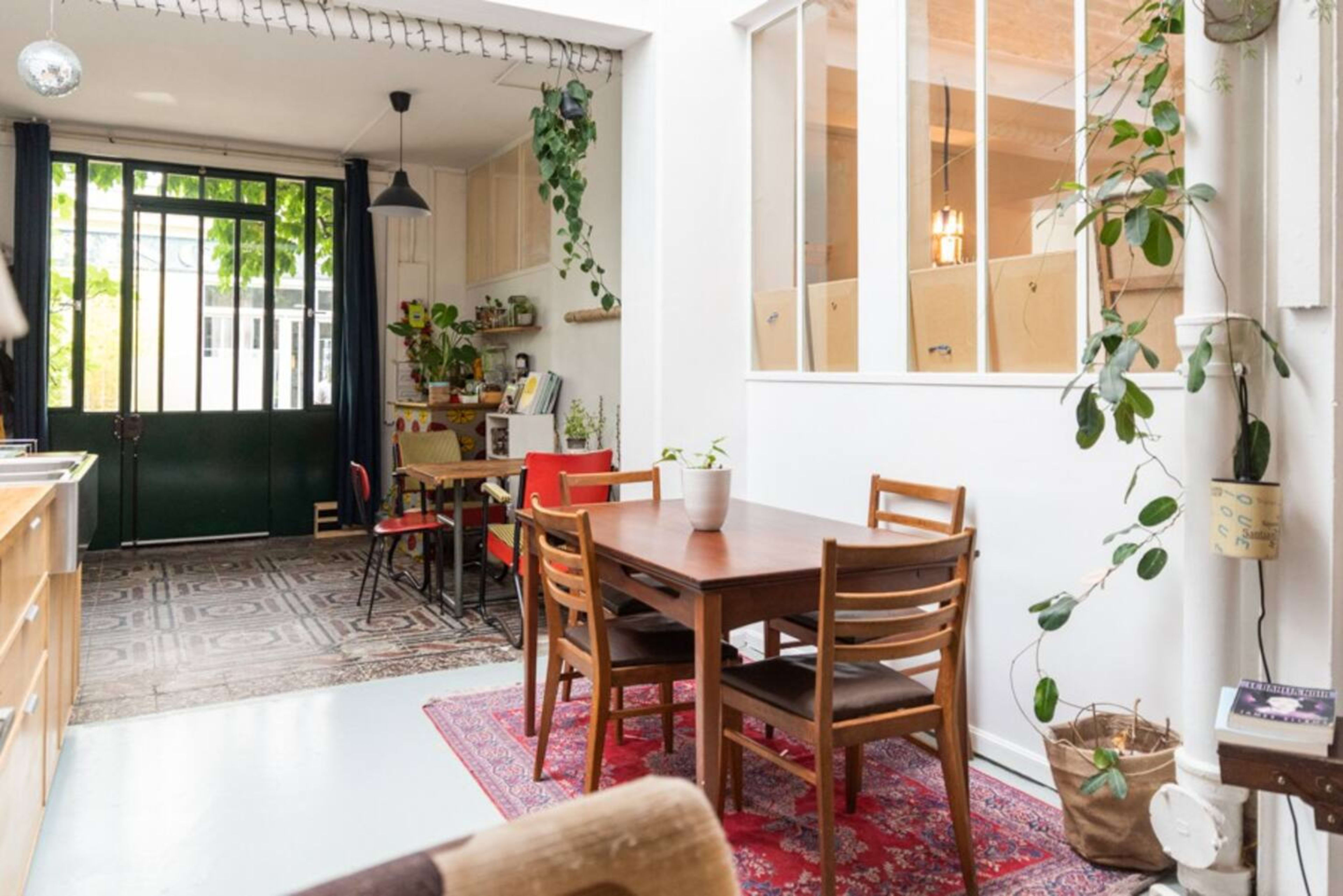A cozy dining area features a wooden table with chairs, surrounded by potted plants and an open view into a brightly lit kitchen.