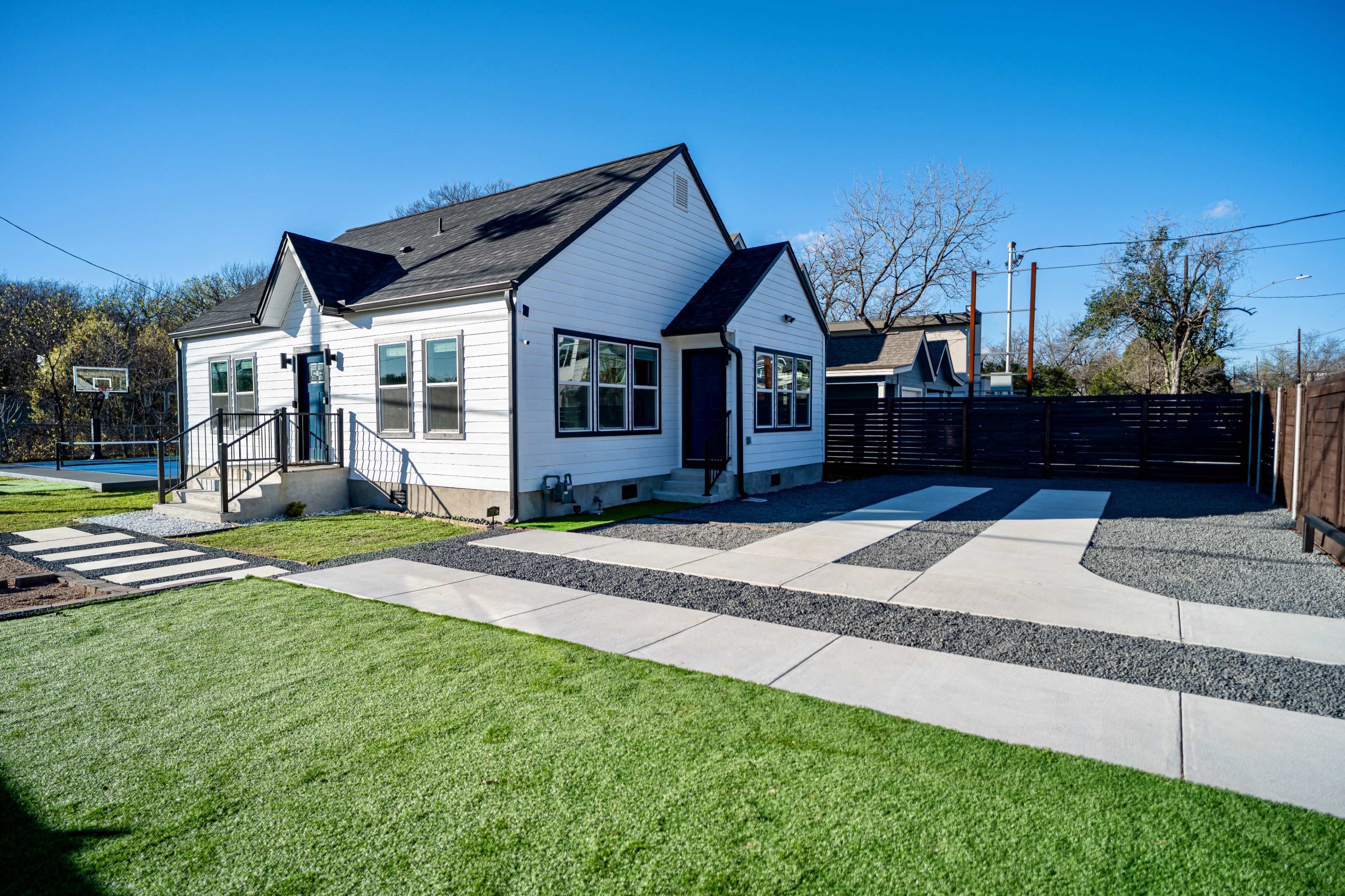 A white house with multiple windows and a blue door sits beside a gravel driveway, surrounded by green grass and a wooden fence.