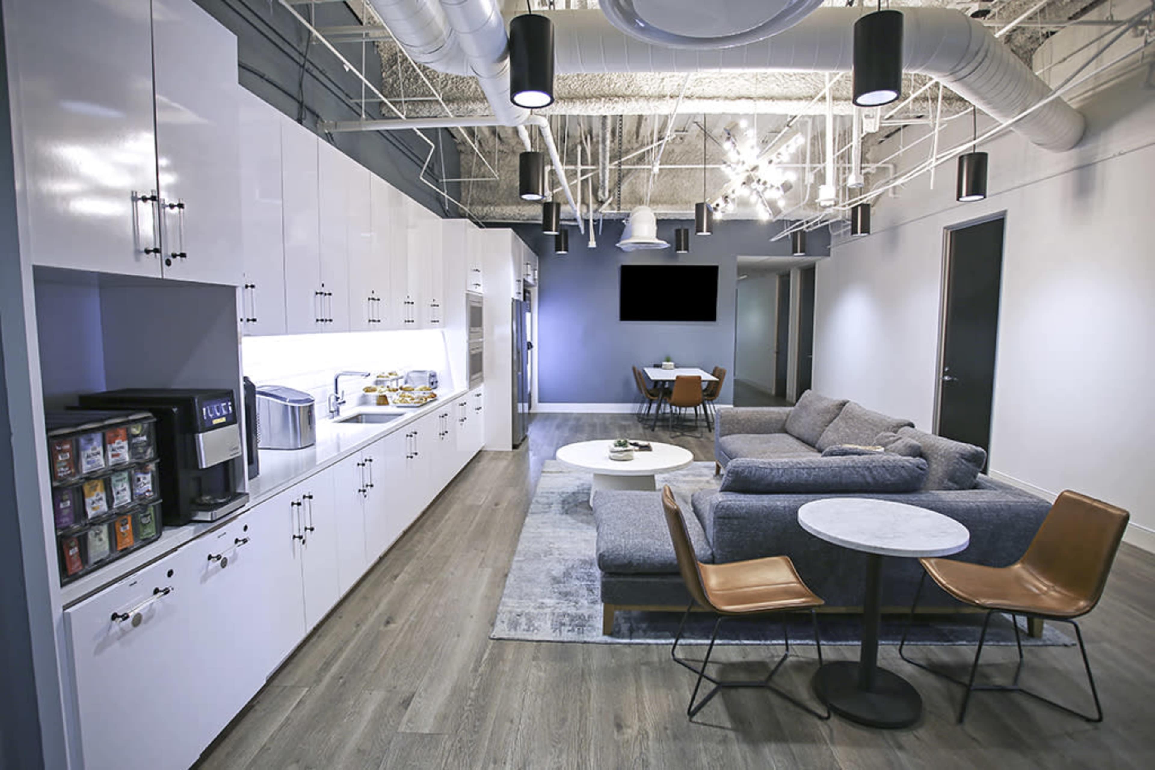 A modern office break room with a lounge area, kitchen cabinets, and a small dining table.