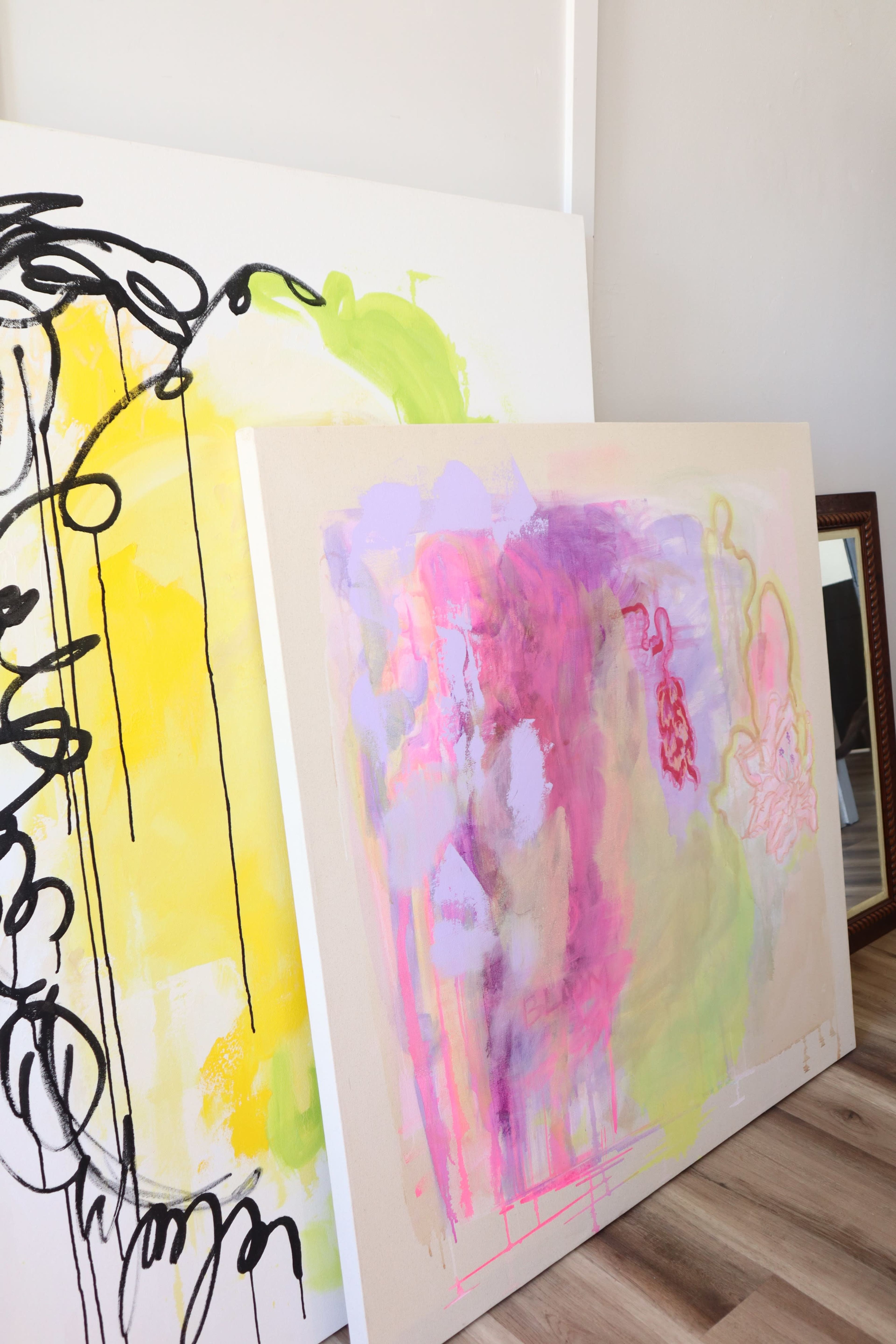 The image features two abstract paintings leaning against a wall, one with vibrant colors and the other in softer hues, alongside a framed mirror.