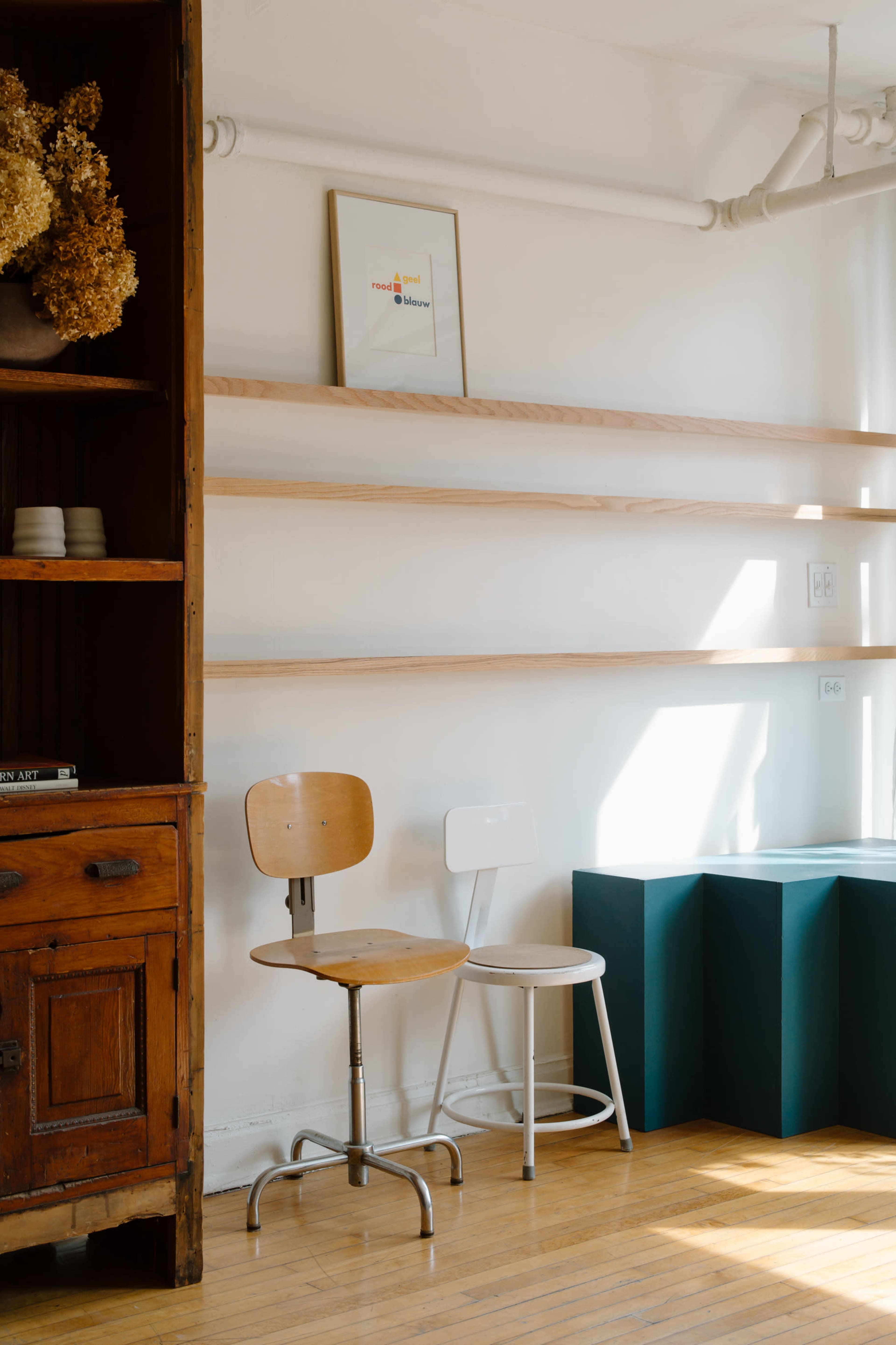 The image shows a minimalist interior featuring two chairs, one wooden and one white, with open wooden shelves against a wall and a decorative cabinet in the corner.
