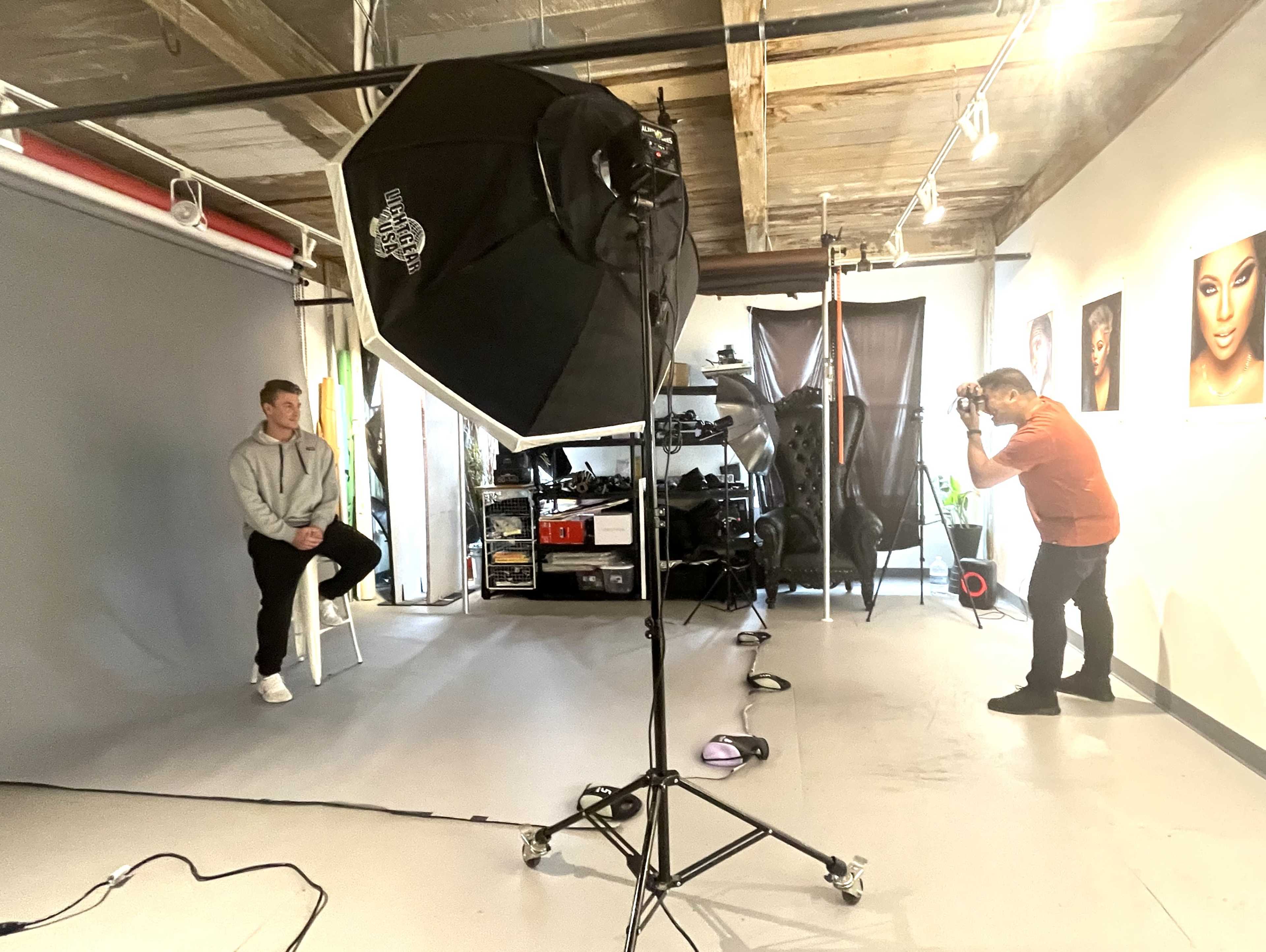 A photographer captures a model sitting on a stool in a studio equipped with lighting and various photography equipment.