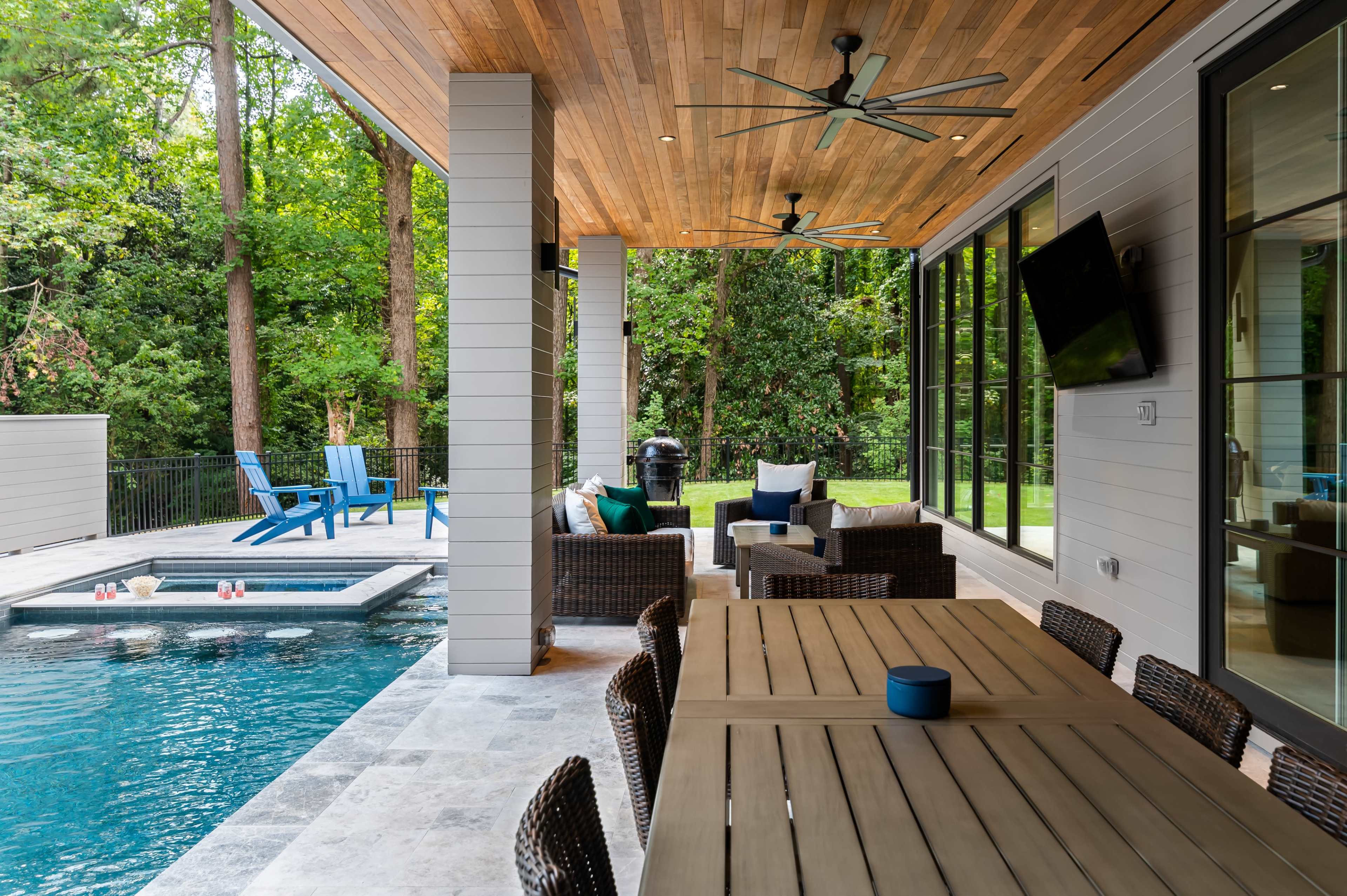 The image shows a modern outdoor patio area with a dining table, seating, and a swimming pool surrounded by greenery.