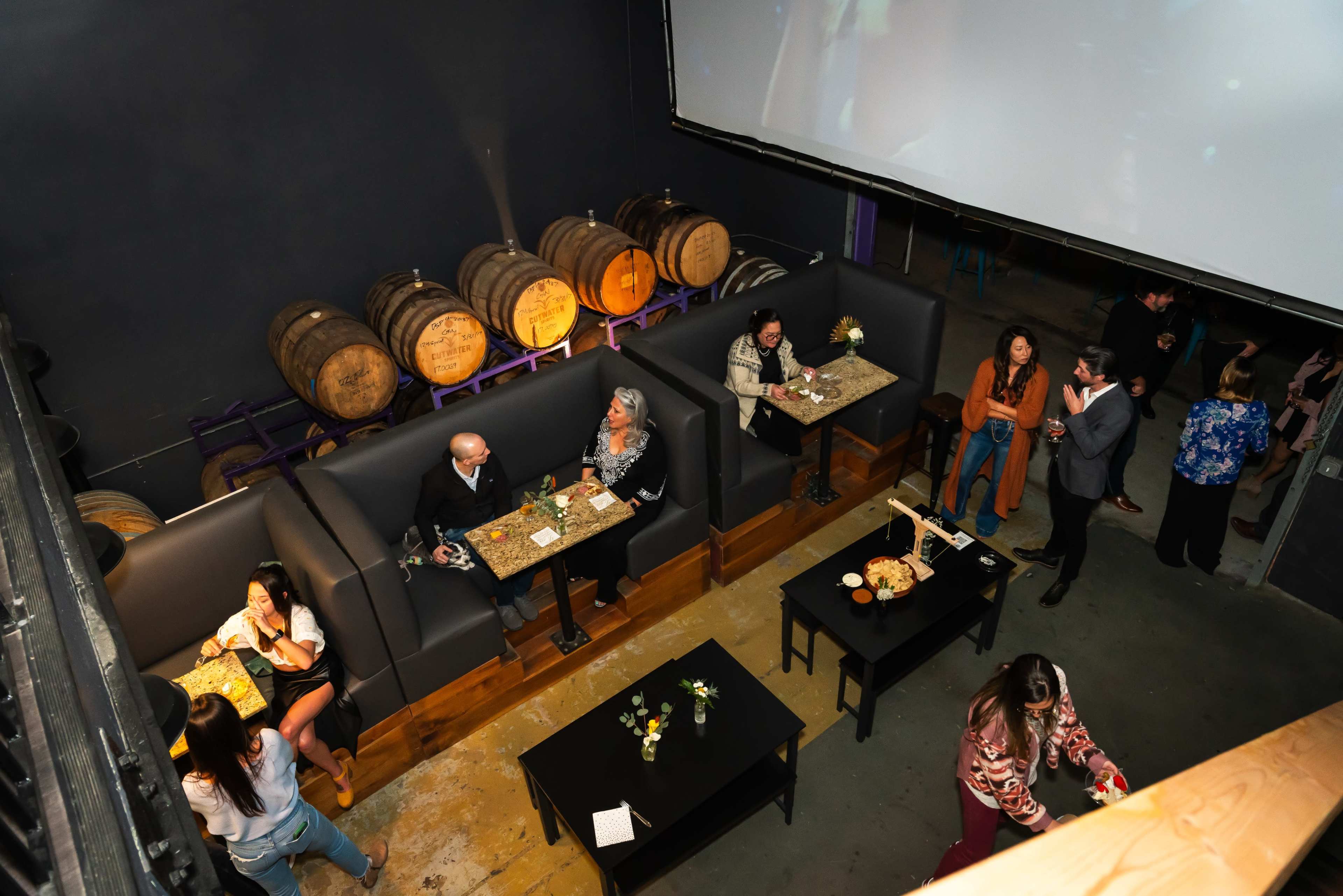 A dimly lit bar setting with patrons seated at tables, surrounded by wooden barrels and a large screen displaying a video.