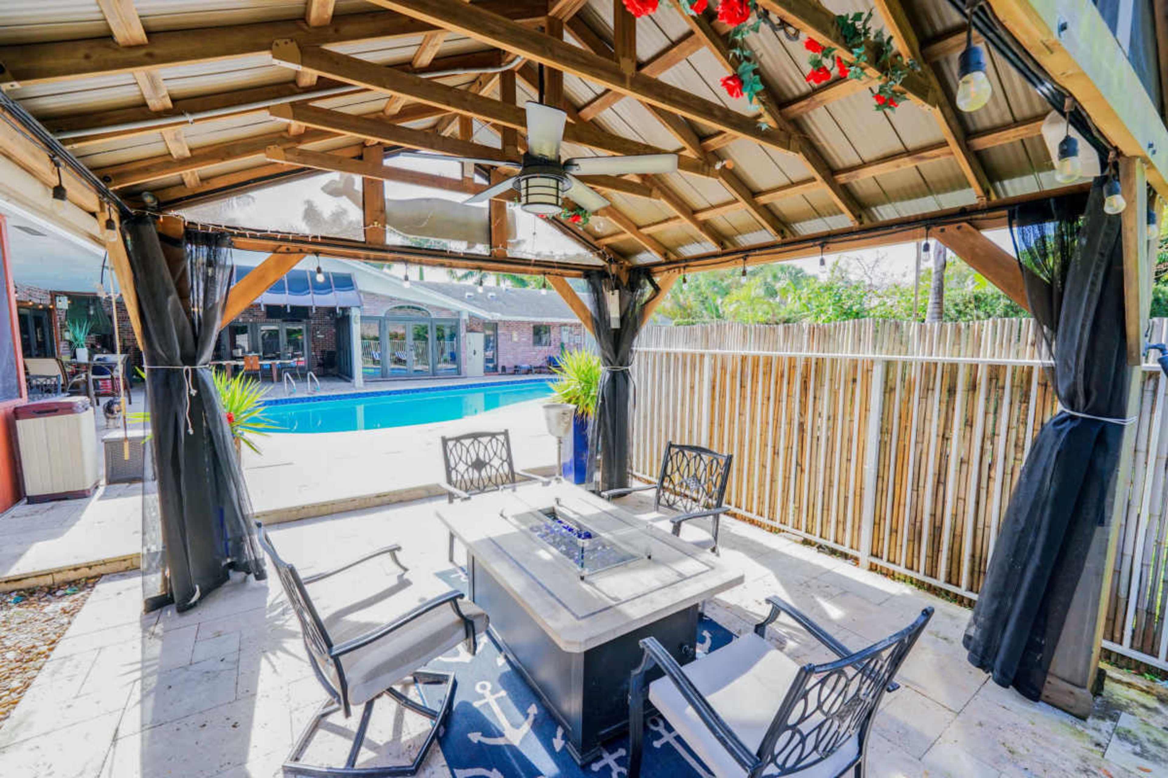A shaded outdoor seating area features a table with a fire pit, surrounded by four chairs, overlooking a swimming pool and fenced yard.
