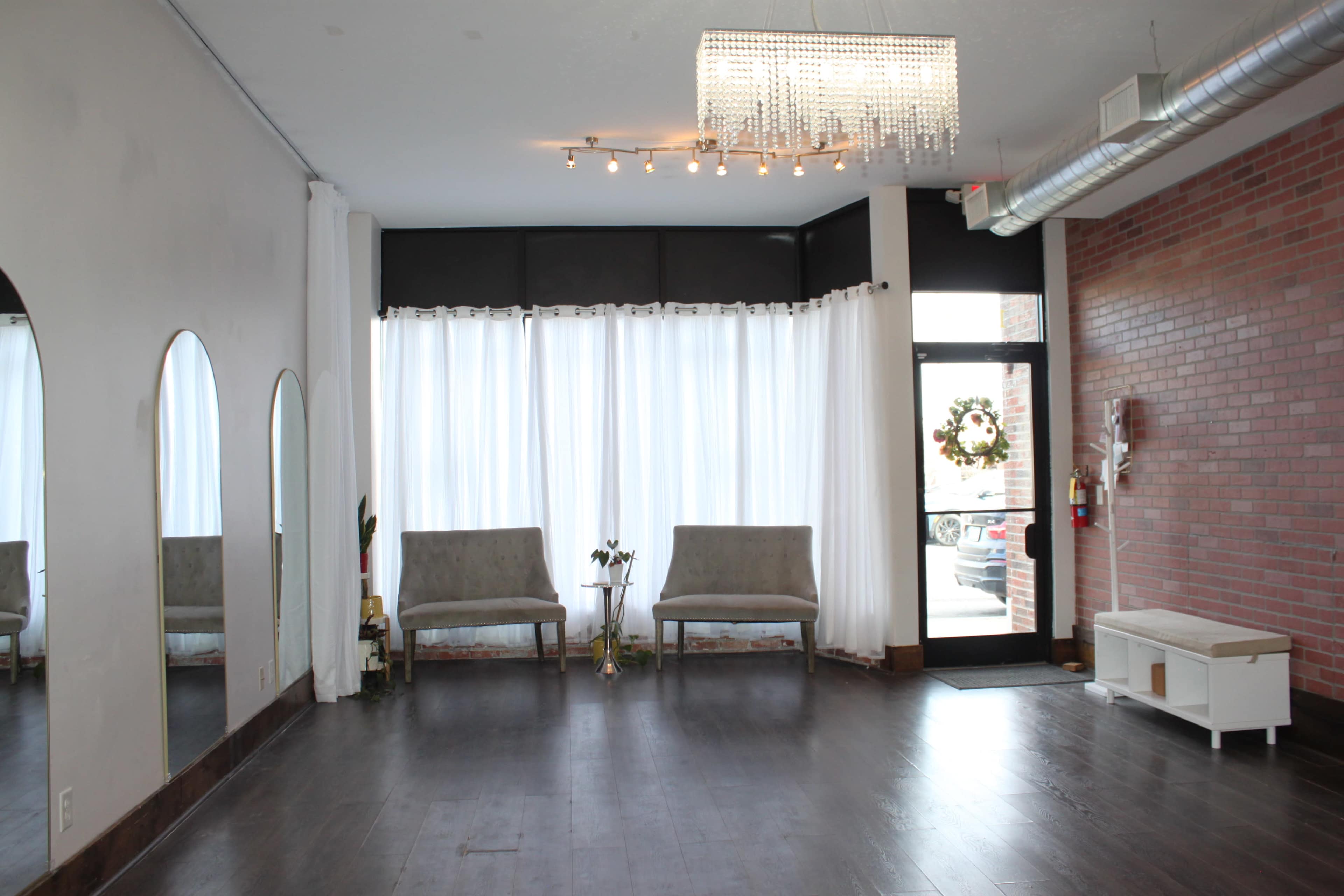 The image shows a spacious interior with two gray chairs, a large window covered by sheer curtains, and a brick wall, all illuminated by a chandelier.