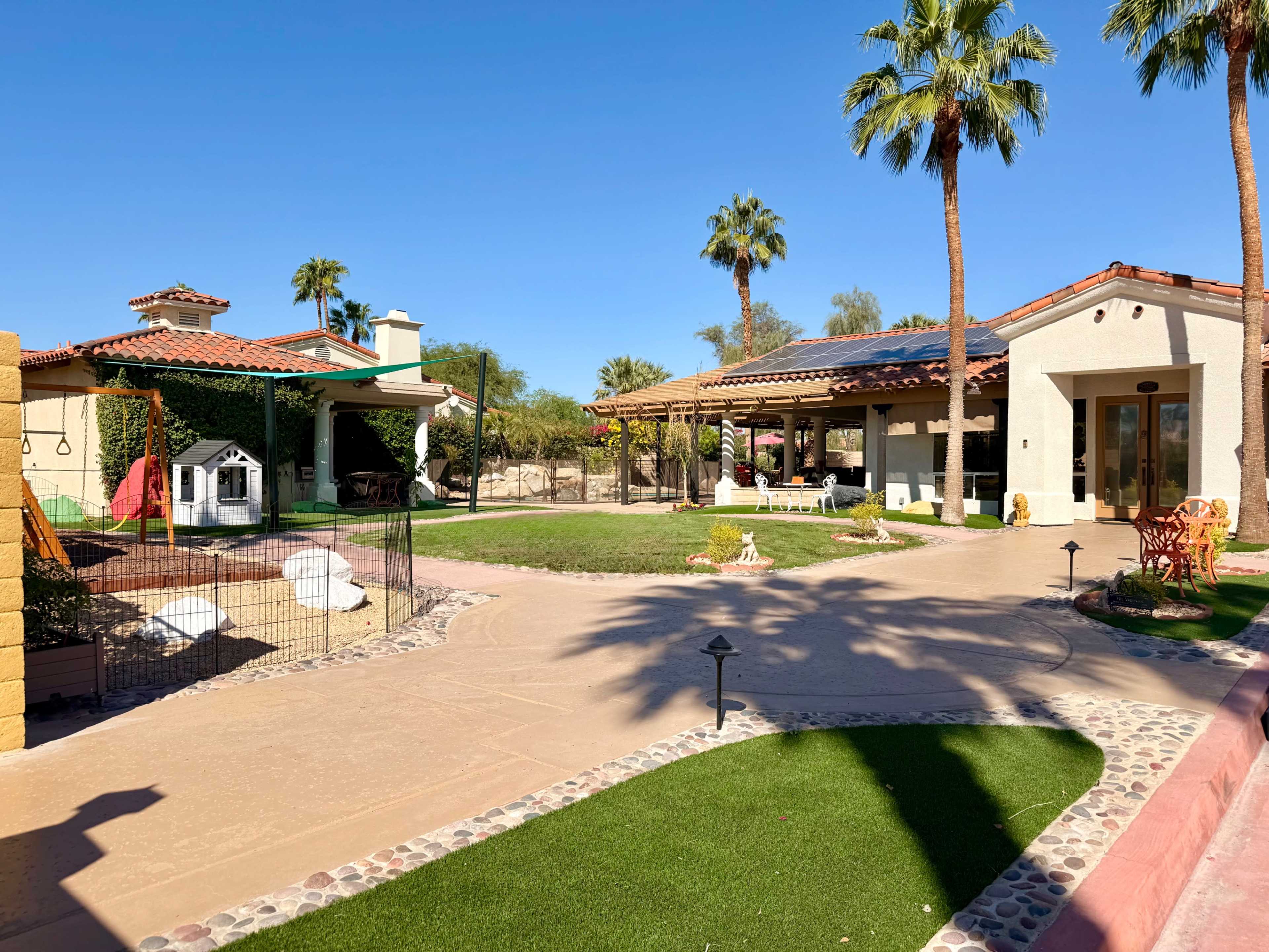 The image shows a landscaped courtyard featuring a single-story house, a play area with a swing set, and several palm trees under a clear blue sky.