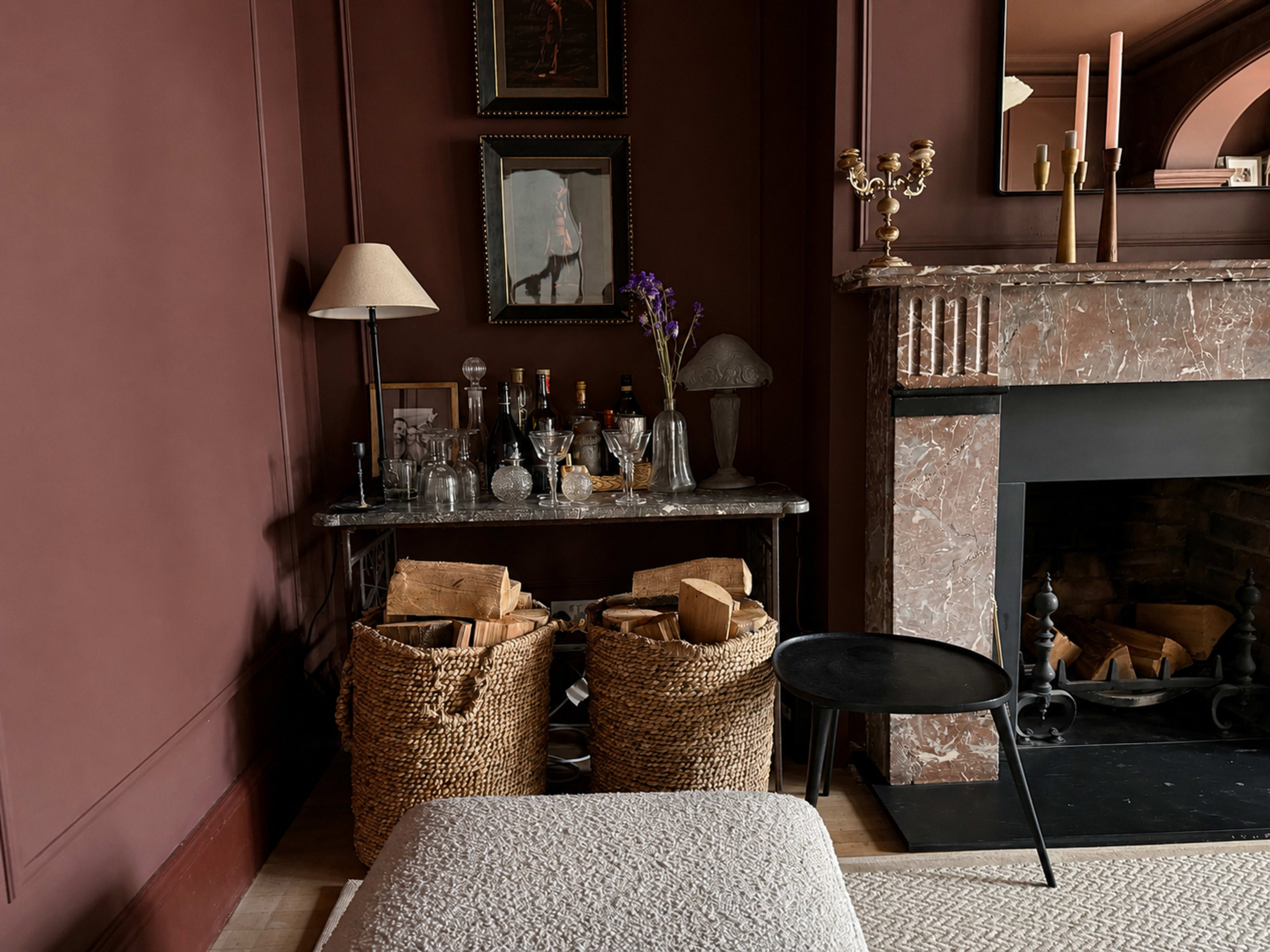 The image shows a cozy interior corner with a marble-topped table featuring glassware and liquor bottles, flanked by woven baskets filled with firewood, and a fireplace with a dark mantel.
