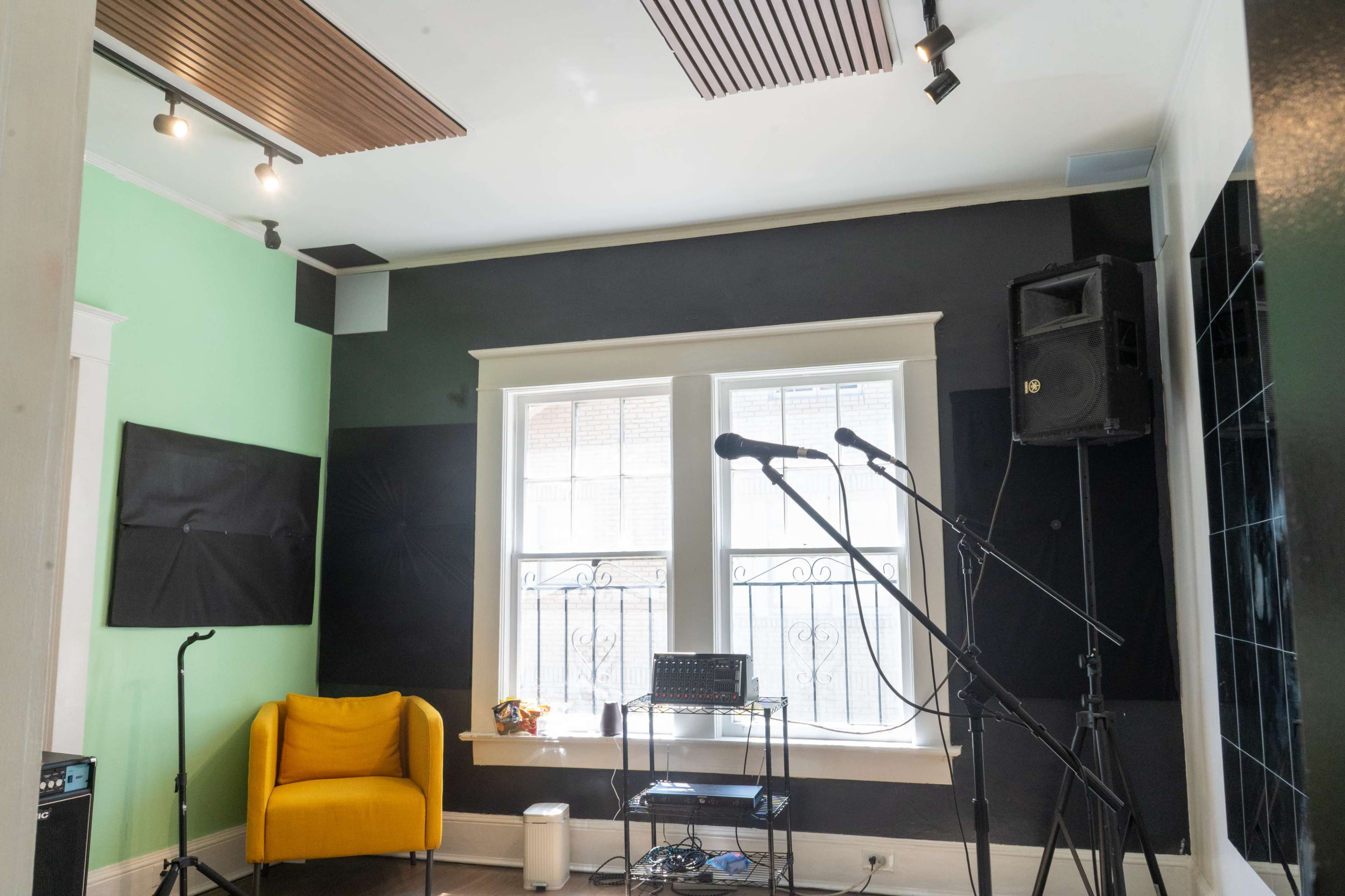 The image shows a music practice room featuring a yellow armchair, two microphones on stands, and sound-absorbing panels on the walls.