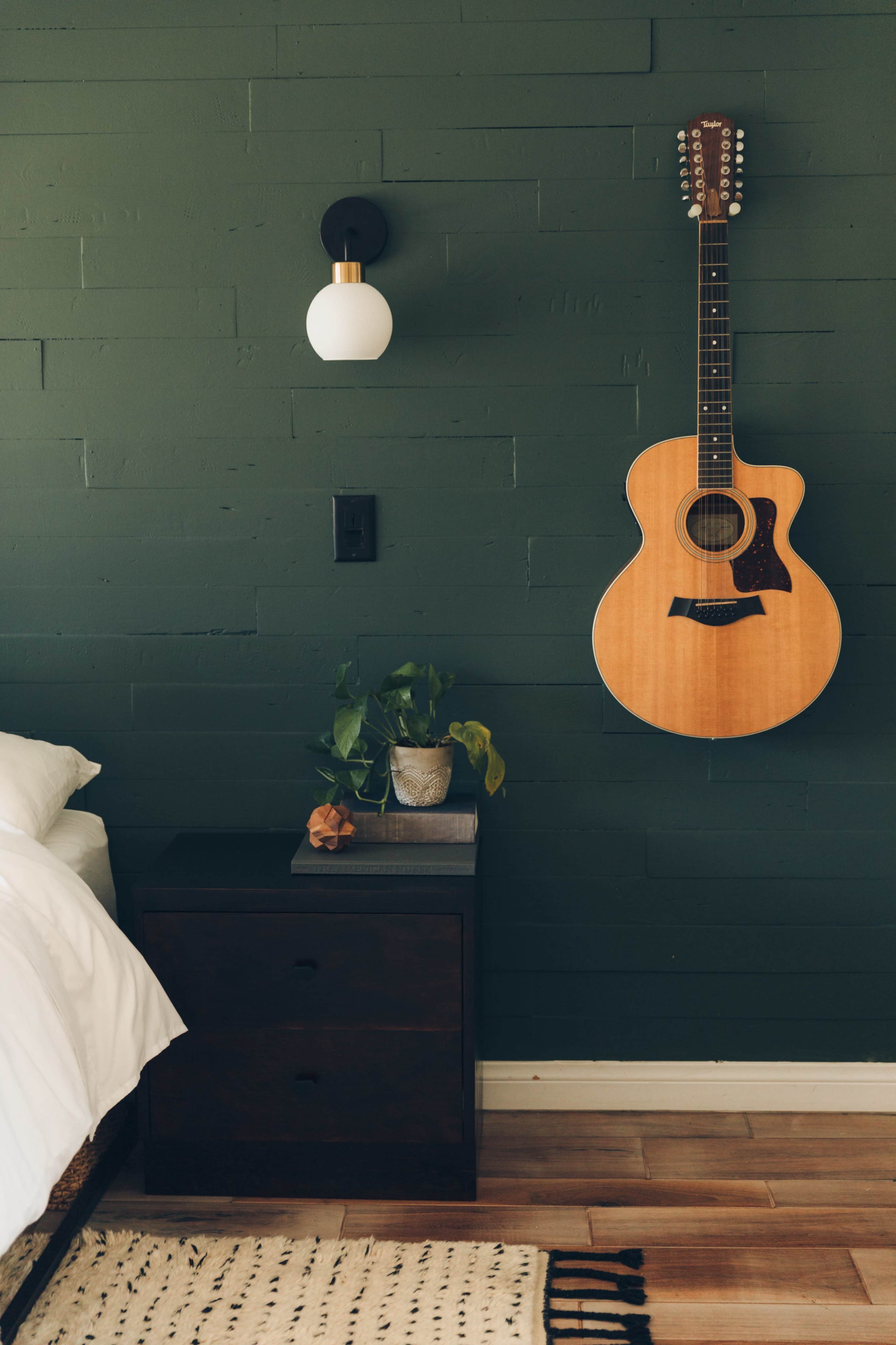 A guitar hangs on a green wall next to a bedside table with a plant on top.