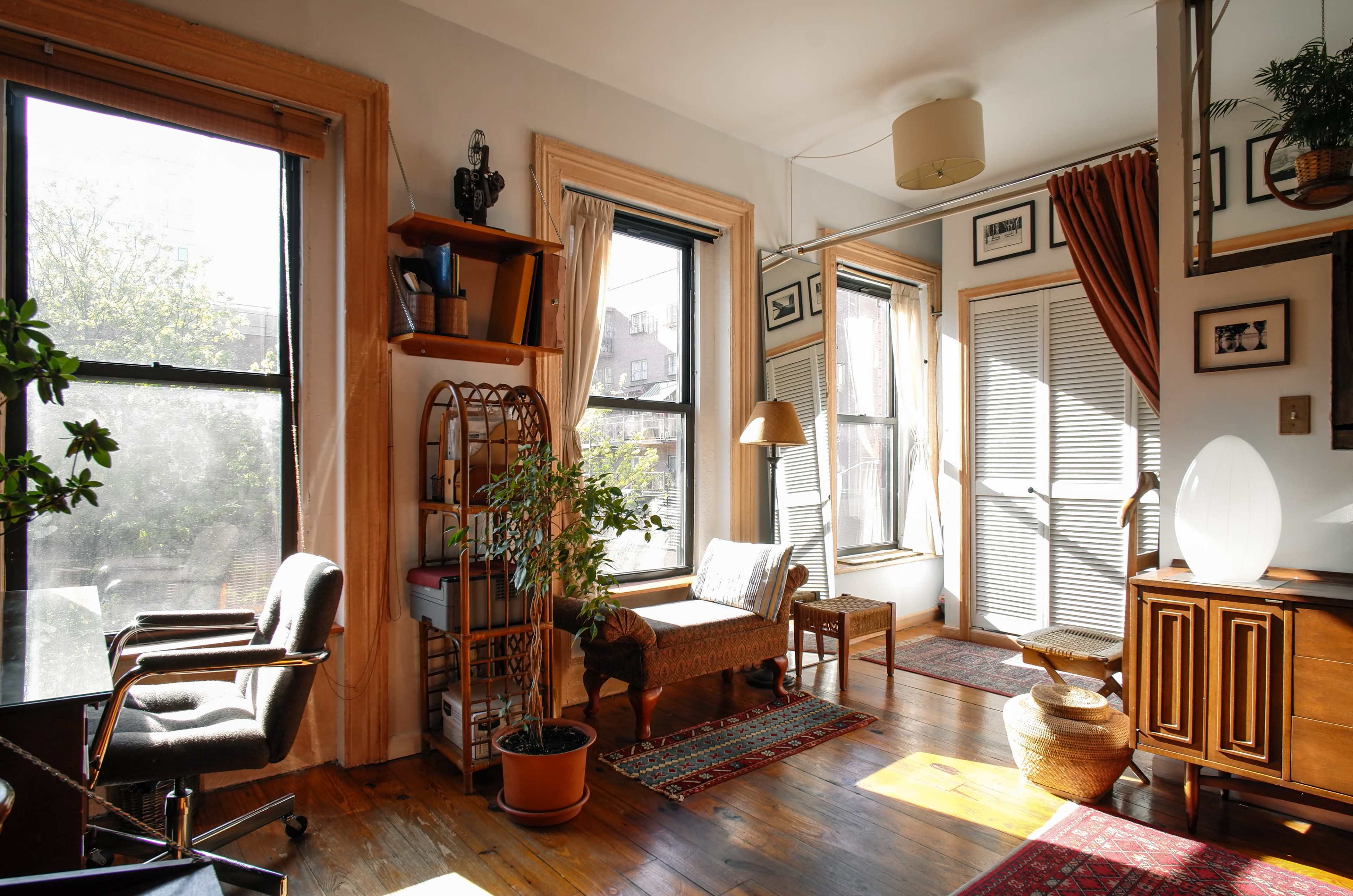 The image depicts a cozy room with large windows, a small seating area, and wooden floors, complemented by plants and a mix of furniture.