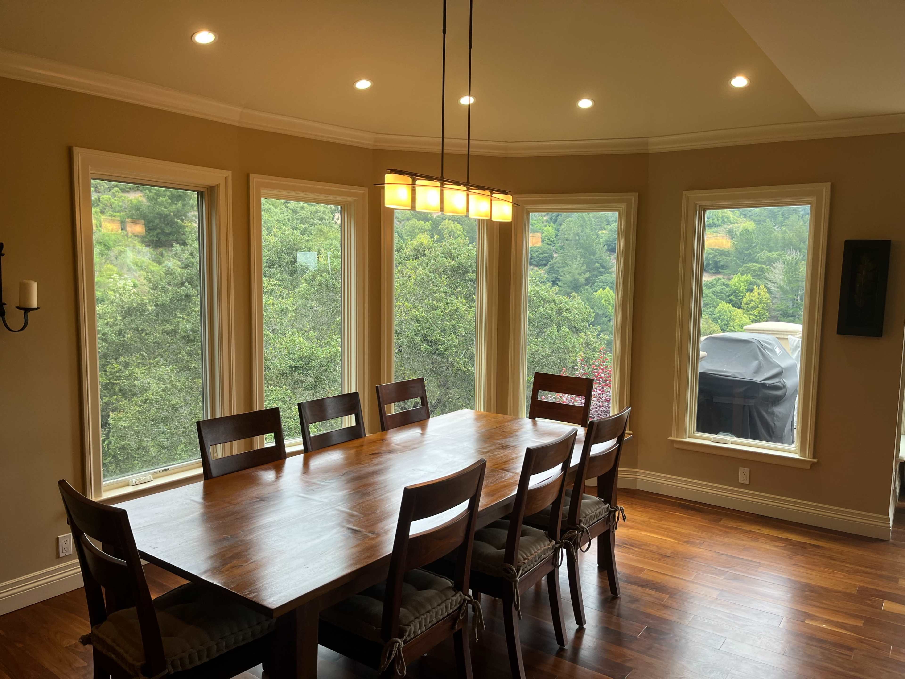 A wooden dining table is positioned in a room with large windows overlooking lush greenery.