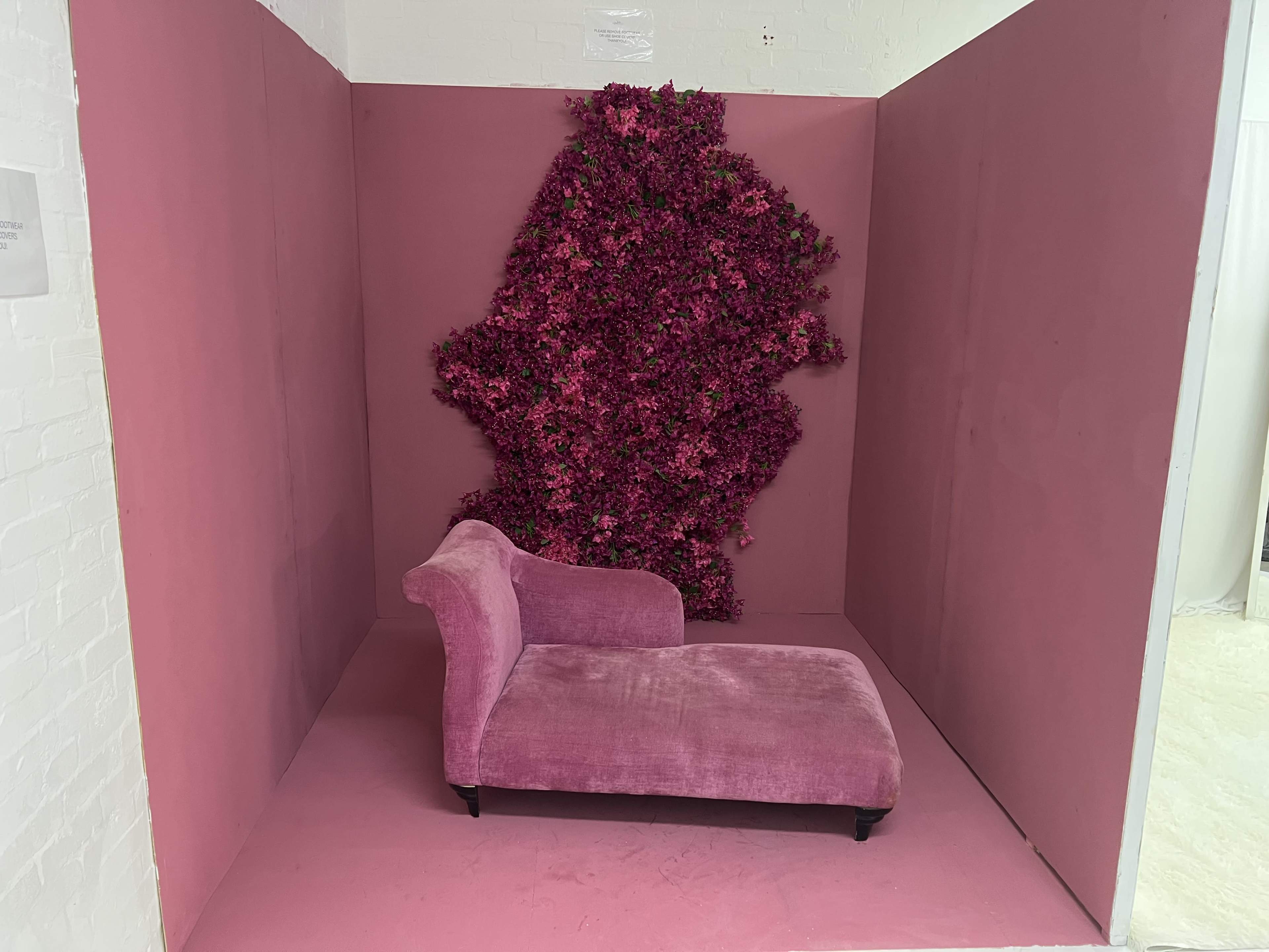 A pink chaise lounge is positioned against a wall covered in vibrant pink floral décor within a similarly colored room.