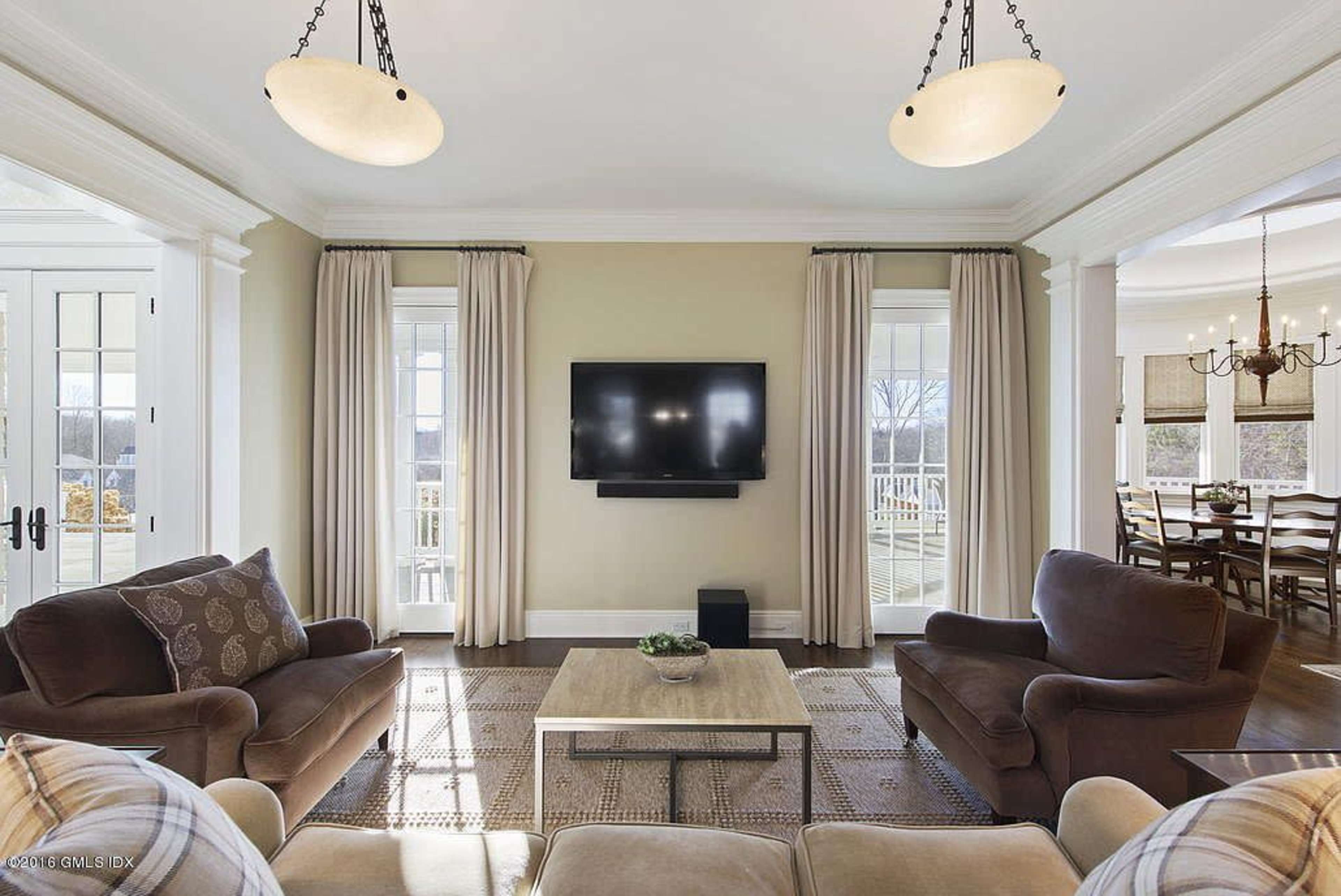 A living room features two brown sofas facing a coffee table, with a mounted television on the wall and large windows flanked by curtains.