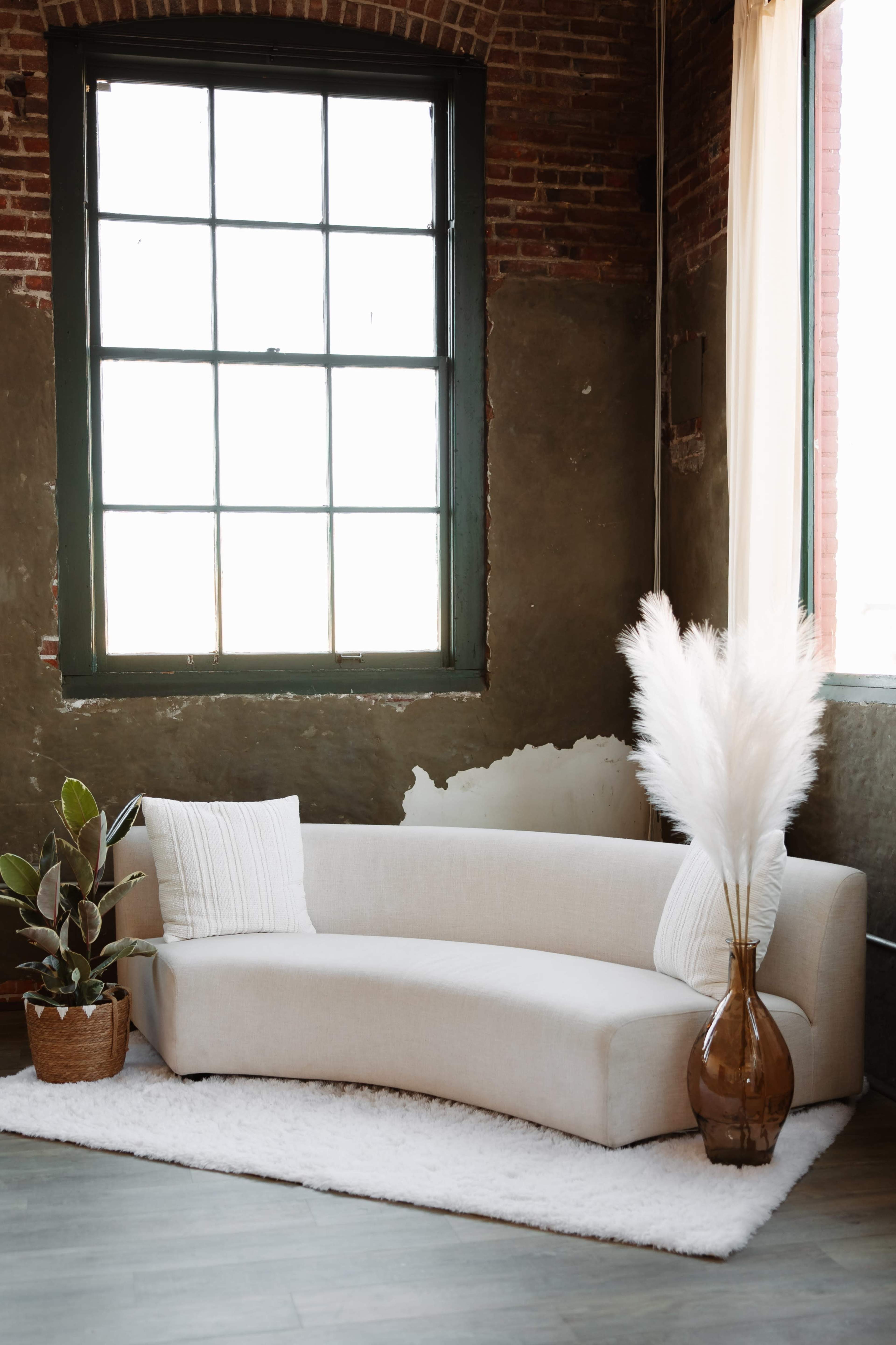 A curved white sofa is positioned near a large window, with a potted plant and a vase of pampas grass on a plush rug in a rustic room with exposed brick walls.