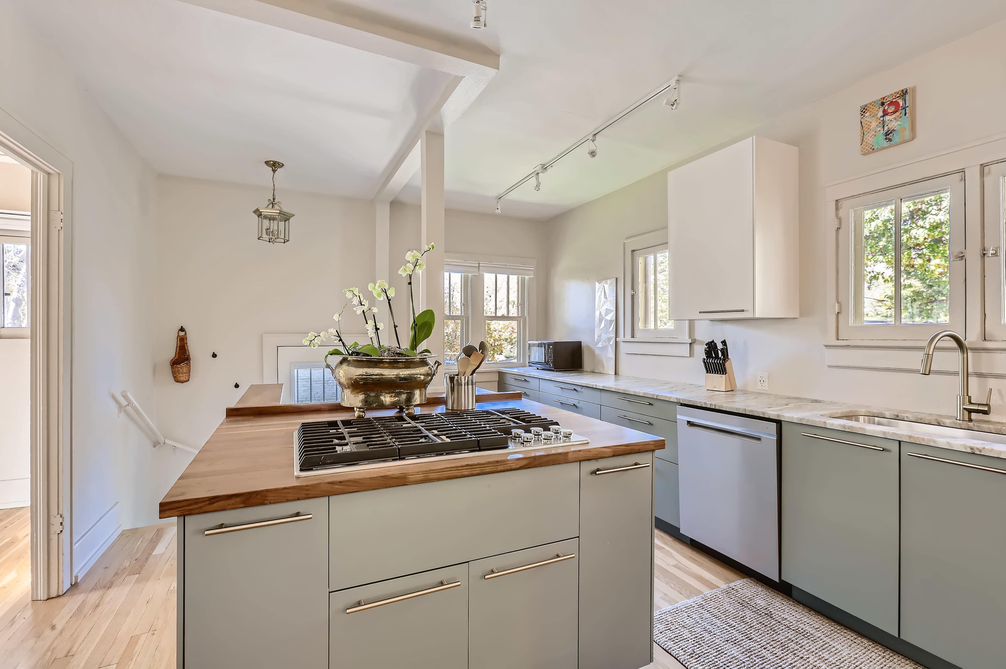 The modern kitchen features a central island with a gas cooktop, light green cabinetry, and large windows that welcome natural light.
